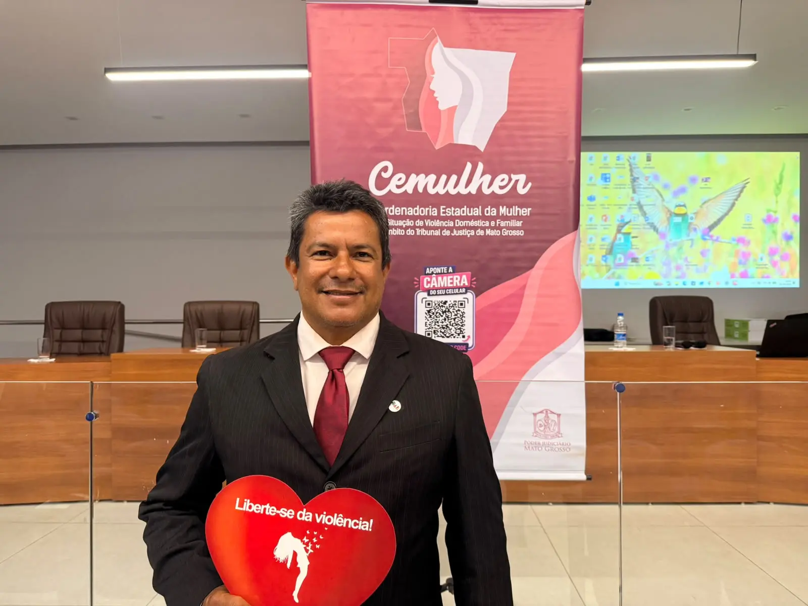 Foto horizontal que mostra o advogado Marcos Miranda da cintura para cima, em pé em frente a um banner da Cemulher, no plenário da Câmara Municipal. Ele é um homem negro, de cabelo grisalho. Usa camisa branca, gravata vermelha, terno preto e segura um panfleto vermelho em formato de coração.