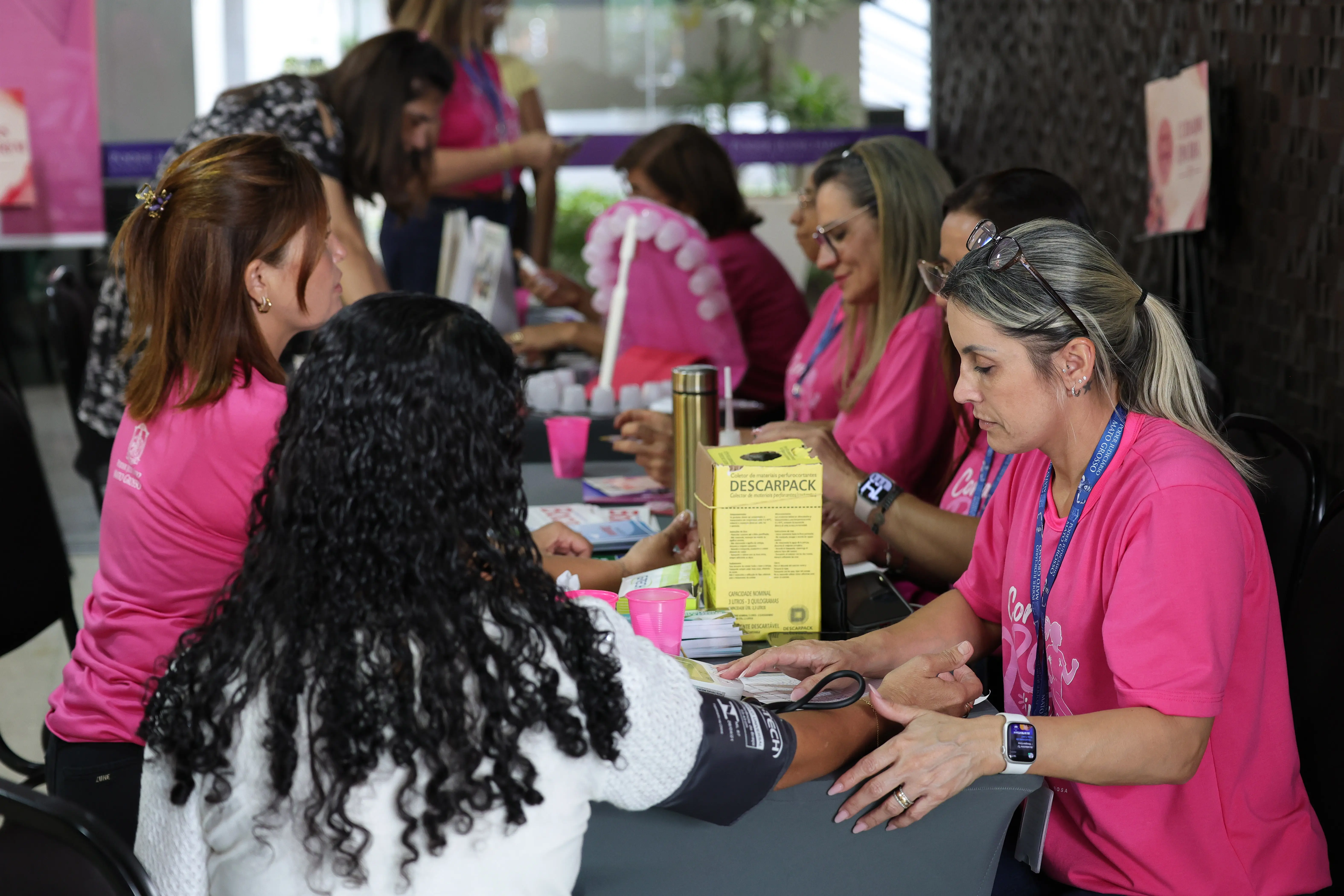 Mulher de cabelos cacheados tem a pressão aferida por servidora de cabelo loiro e camiseta rosa, em atendimento de saúde do Outubro Rosa; ao fundo, outras profissionais atendem.
