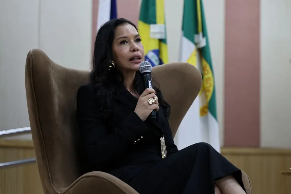 Foto horizontal em plano médio que mostra a promotora de justiça Alessandra Godoi sentada em uma poltrona marrom, falando ao microfone. Ela é uma mulher de pele parda, cabelos pretos e longos, usando saia e blazer pretos.