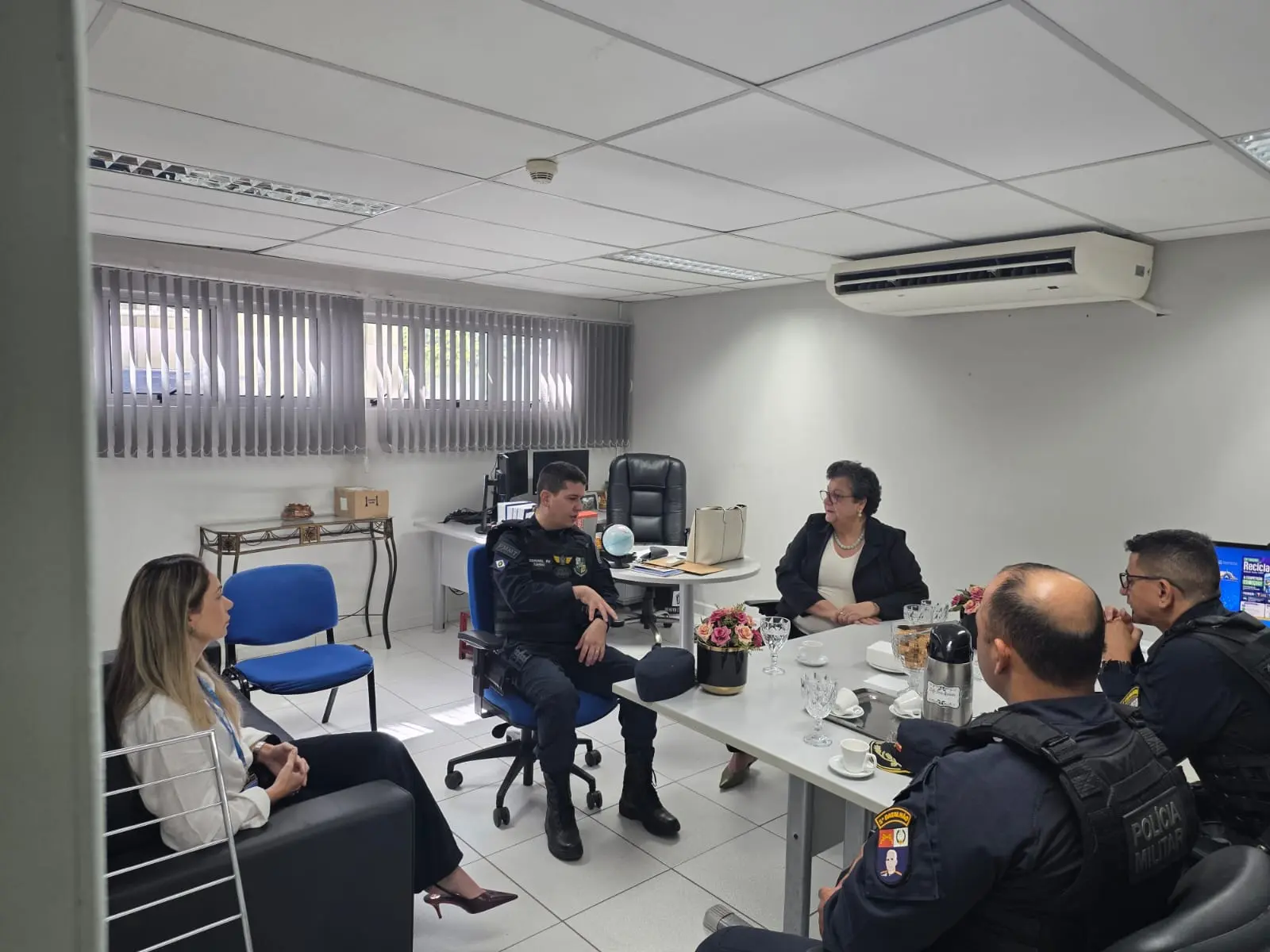 Reunião em escritório sobre violência contra a mulher. Uma mulher loira, um policial sentado ao centro, uma mulher de preto ao fundo e dois policiais de costas conversam ao redor de mesas.