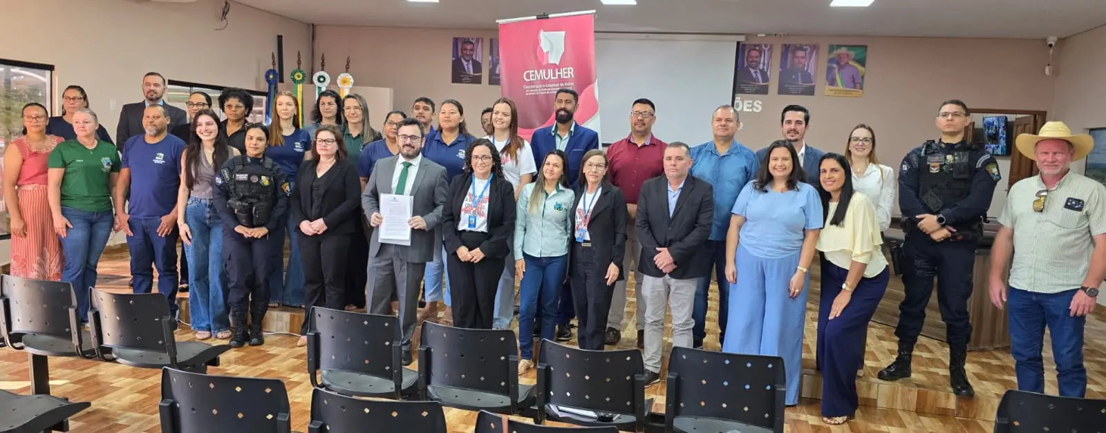 Grande grupo de homens e mulheres posa de pé em um auditório com cadeiras vazias em primeiro plano. Um homem ao centro segura um documento. Ao fundo, um banner vermelho escrito 