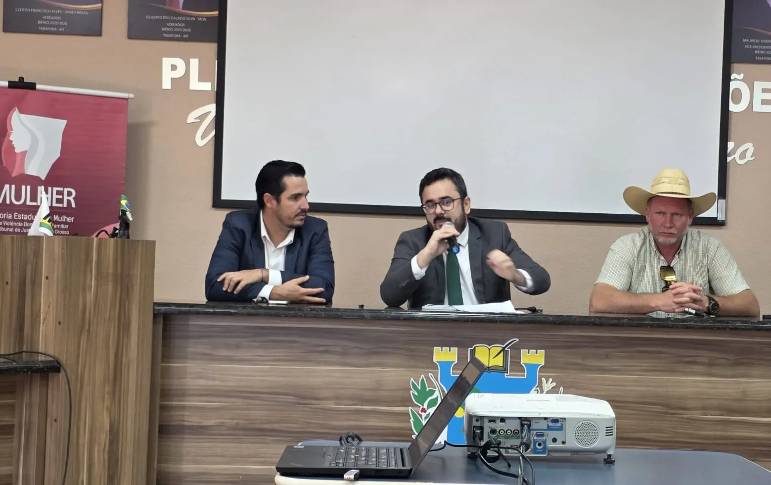 Três homens sentados à mesa de um plenário. O do centro, de terno e óculos, fala ao microfone. À direita, um homem usa chapéu de palha. À frente, há um notebook e um projetor.