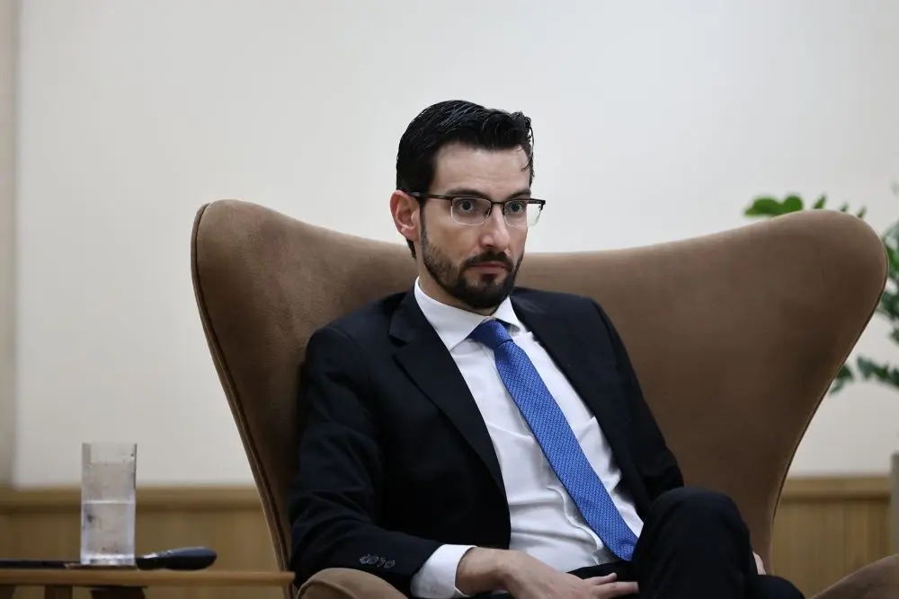 Foto horizontal em plano médio que mostra o promotor de justiça Luiz Fernando Rossi sentado em uma poltrona marrom. Ele é um homem branco, magro, alto, de olhos e cabelos pretos, usando camisa branca, gravata azul e terno preto.