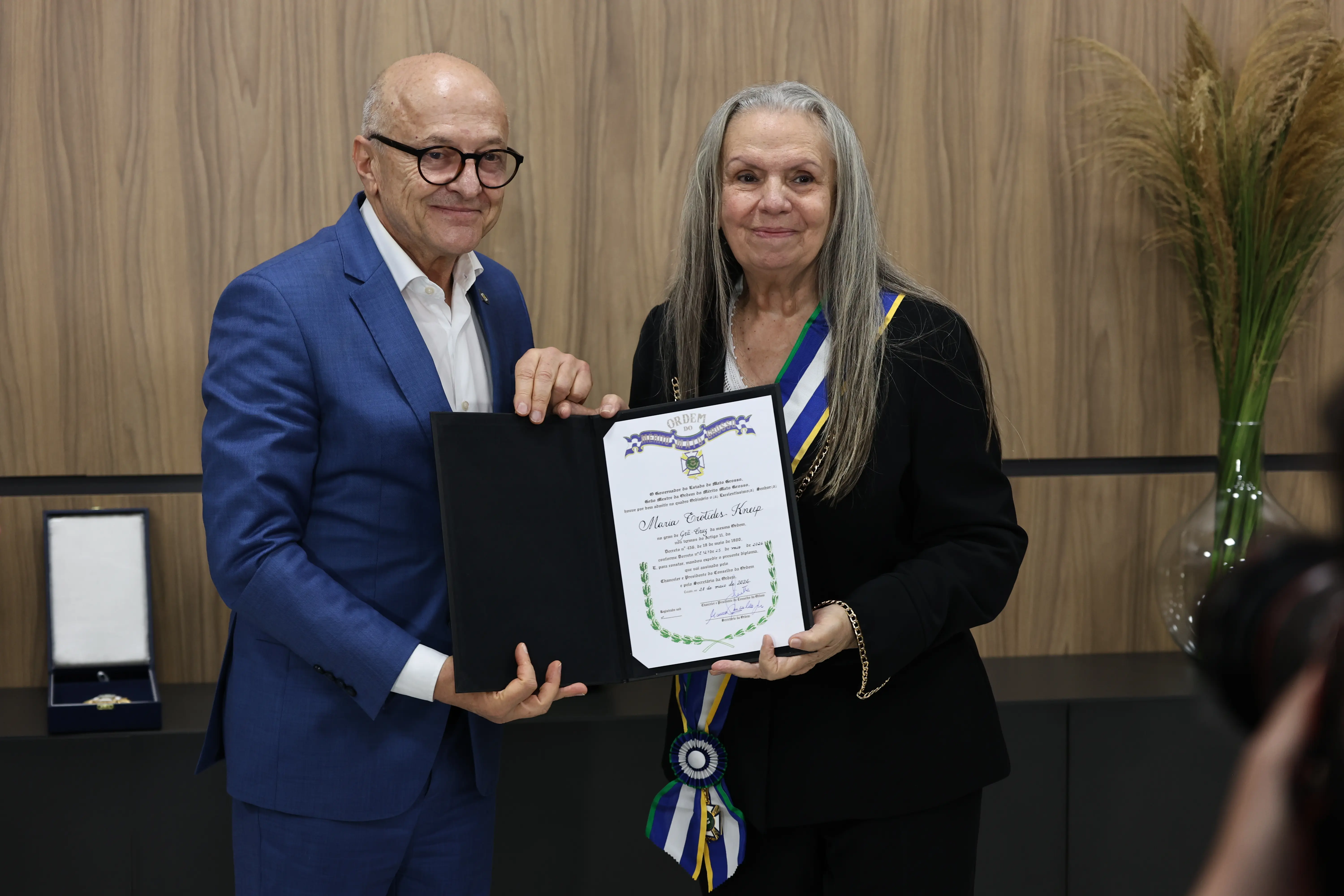 Um homem de terno azul e óculos, e uma mulher idosa de cabelos longos e grisalhos seguram juntos um diploma aberto. Ela usa uma faixa transversal azul, verde e branca. Fundo amadeirado.