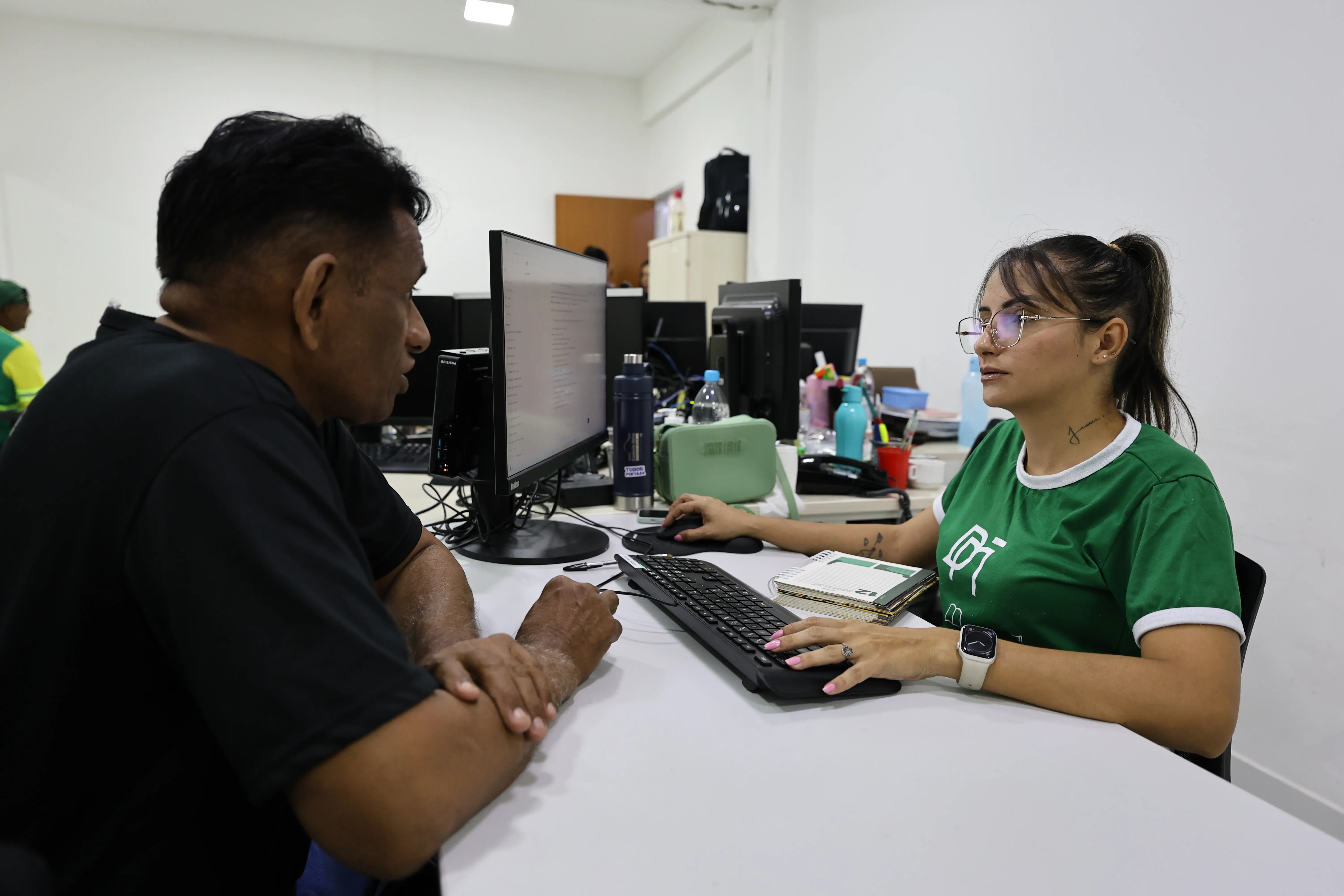 Mulher de camiseta verde e óculos atende homem de camiseta preta em mesa com computador. Ela usa teclado e mouse enquanto conversam em um ambiente de escritório iluminado.
