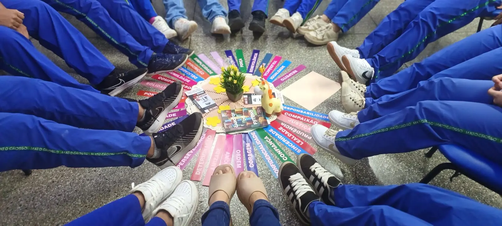 Visão de cima de pernas e pés de estudantes com calças azuis sentados em círculo. No centro, fitas coloridas com palavras de valores apontam para um pequeno vaso de flores e uma girafa de brinquedo.