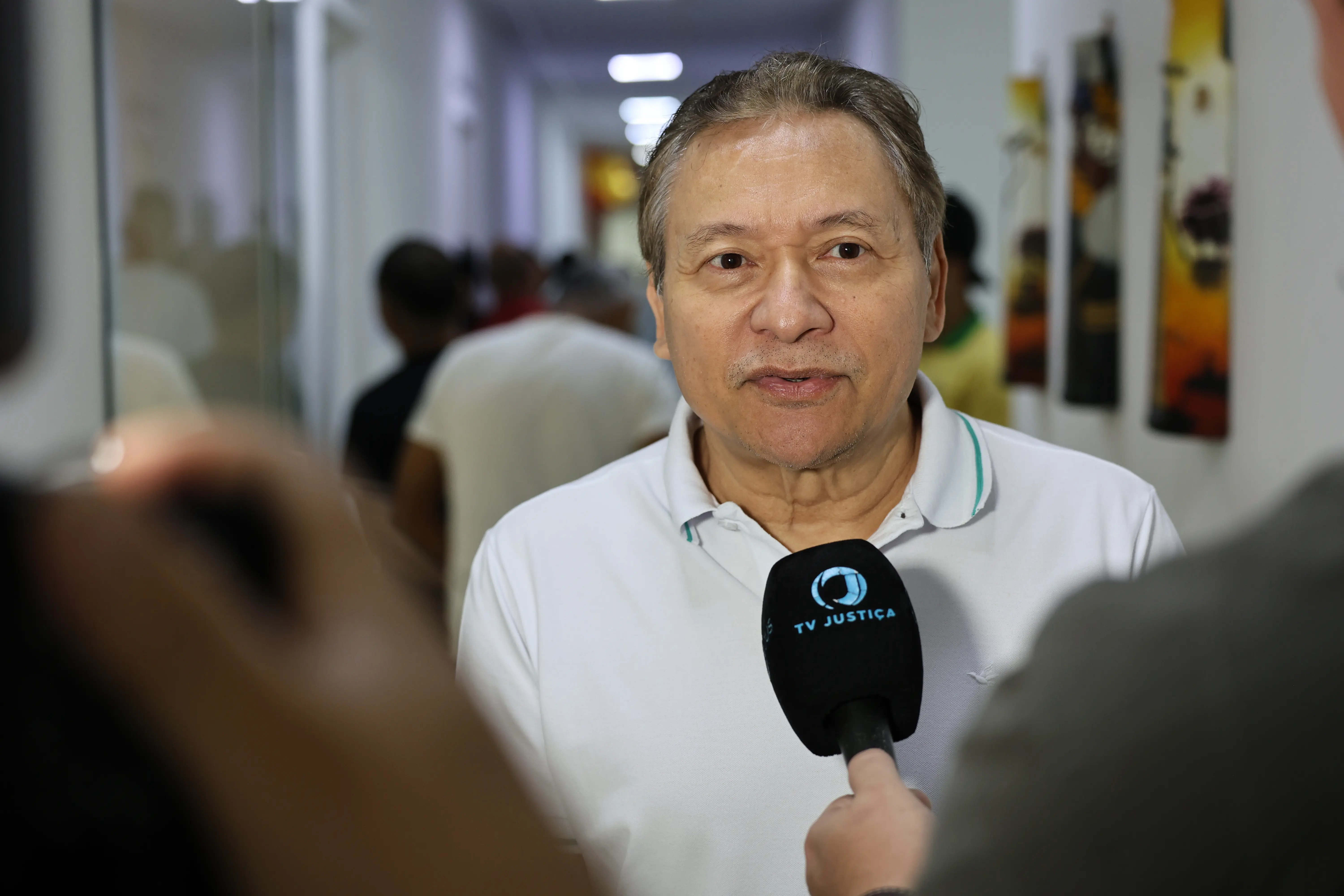 Homem de traços orientais e cabelos grisalhos veste camisa polo branca e fala para um microfone da TV Justiça em um corredor iluminado com pessoas ao fundo.
