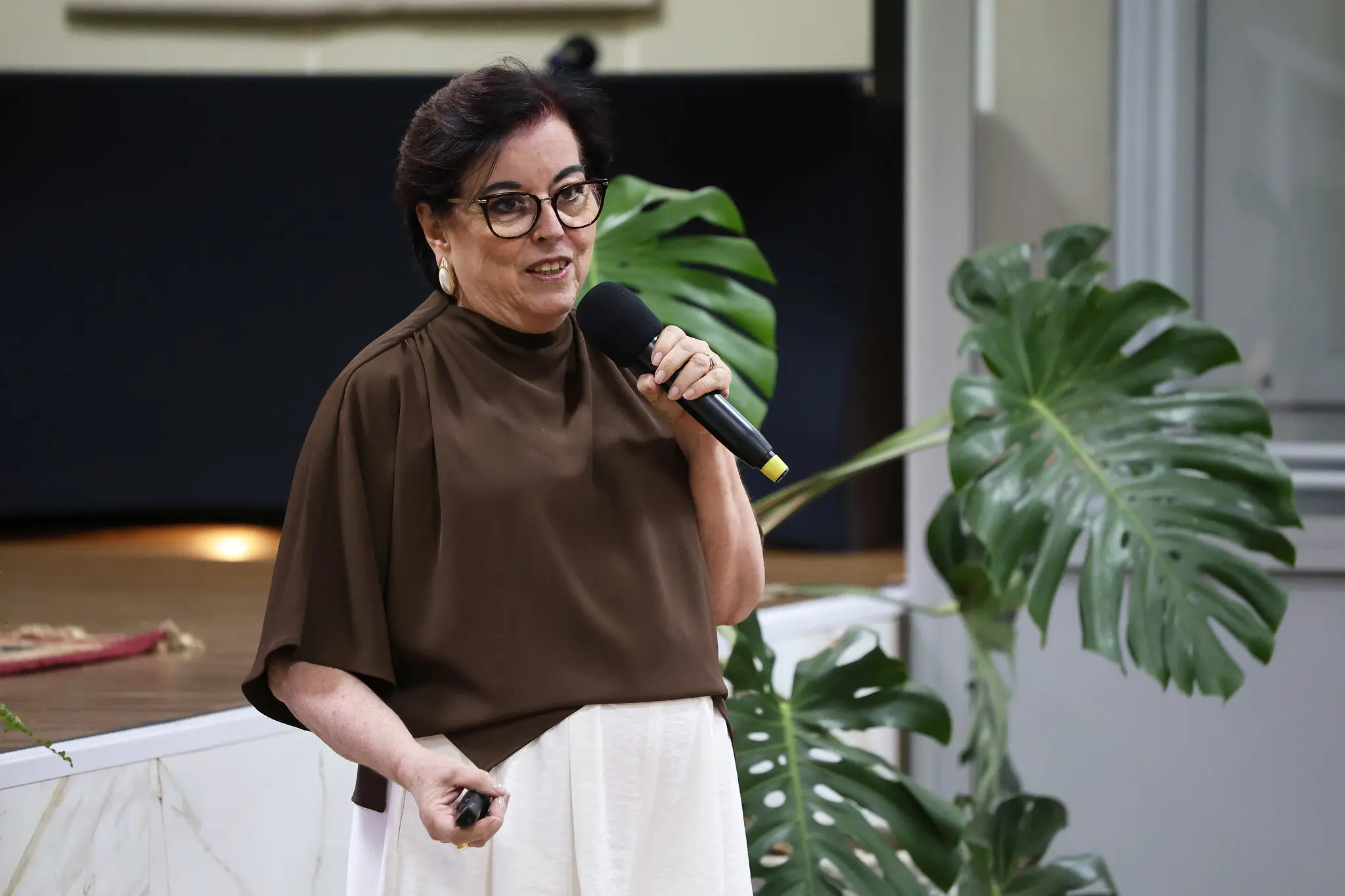 Palestrante fala ao microfone em auditório, usando blusa marrom e saia clara. Planta ornamental está ao lado. Fundo com palco elevado e painel escuro.