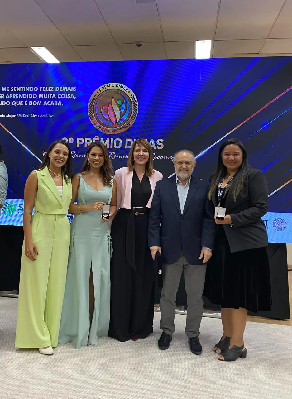 Cinco pessoas posam sorrindo em frente a um telão do Prêmio Dimas. As mulheres usam vestidos e ternos elegantes, algumas segurando medalhas. Um homem de blazer azul está ao centro. Ambiente interno e iluminado.