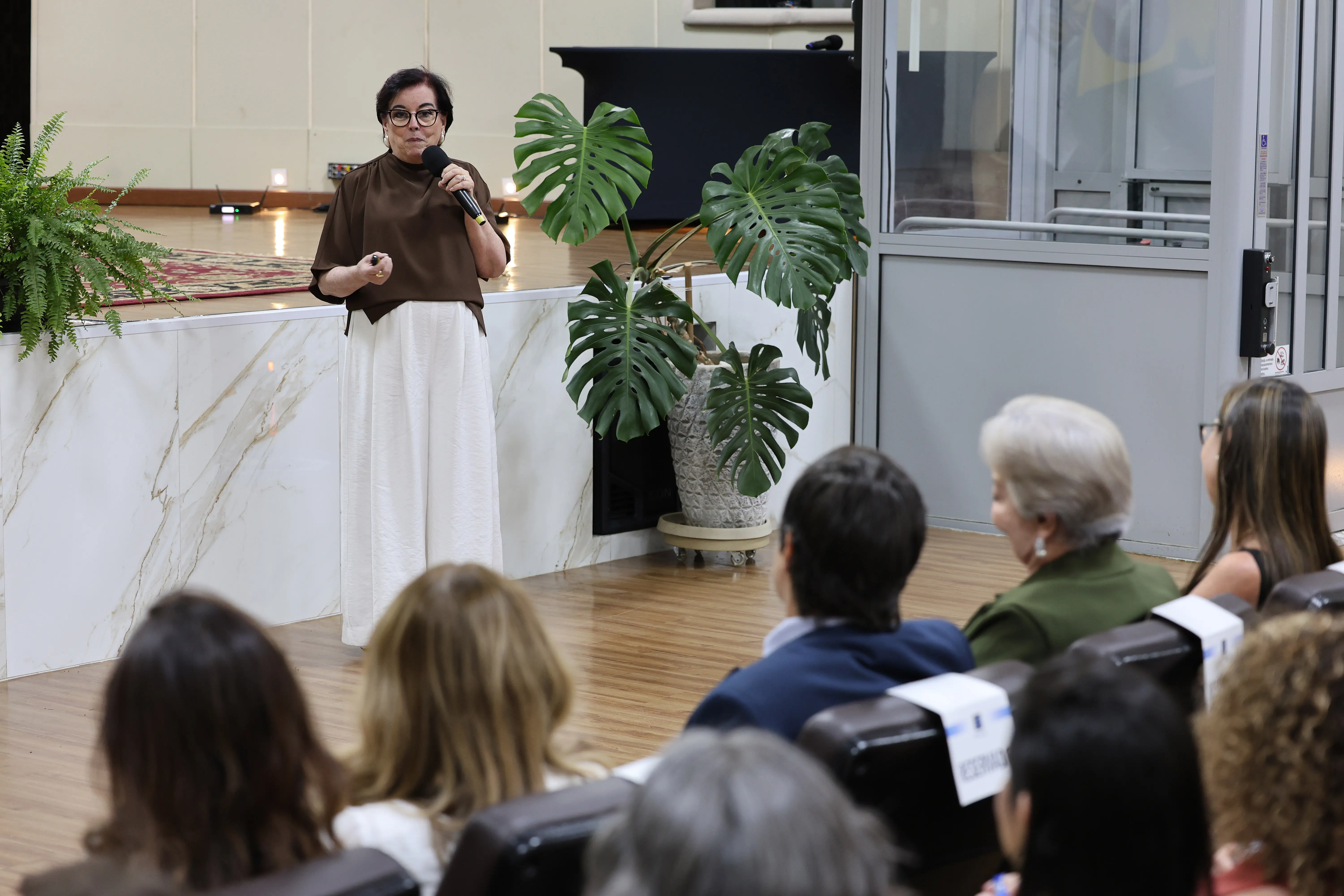 Palestrante segura microfone enquanto fala a um público sentado em auditório. Ela usa blusa marrom e saia clara. Ao fundo, palco com tapete, plantas decorativas e painel de mármore claro.