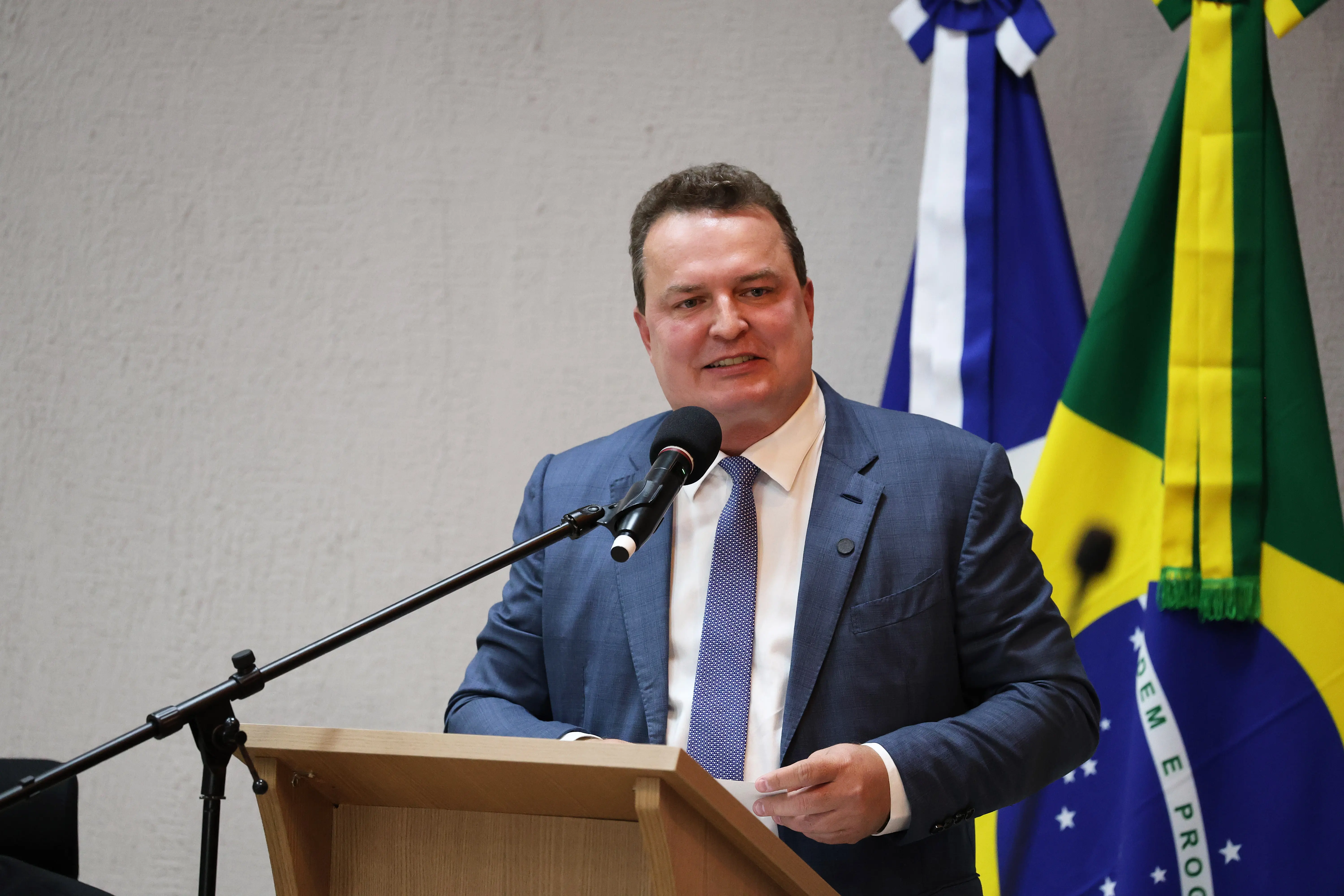 Presidente da Assembleia Legislativa de Mato Grosso, deputado Max Russi, em discurso na tribuna do Tribunal de Justiça. Ele é um homem alto, branco, de olhos verdes, cabelos castanhos claros, usando camisa branca, gravata lilás e terno azul.