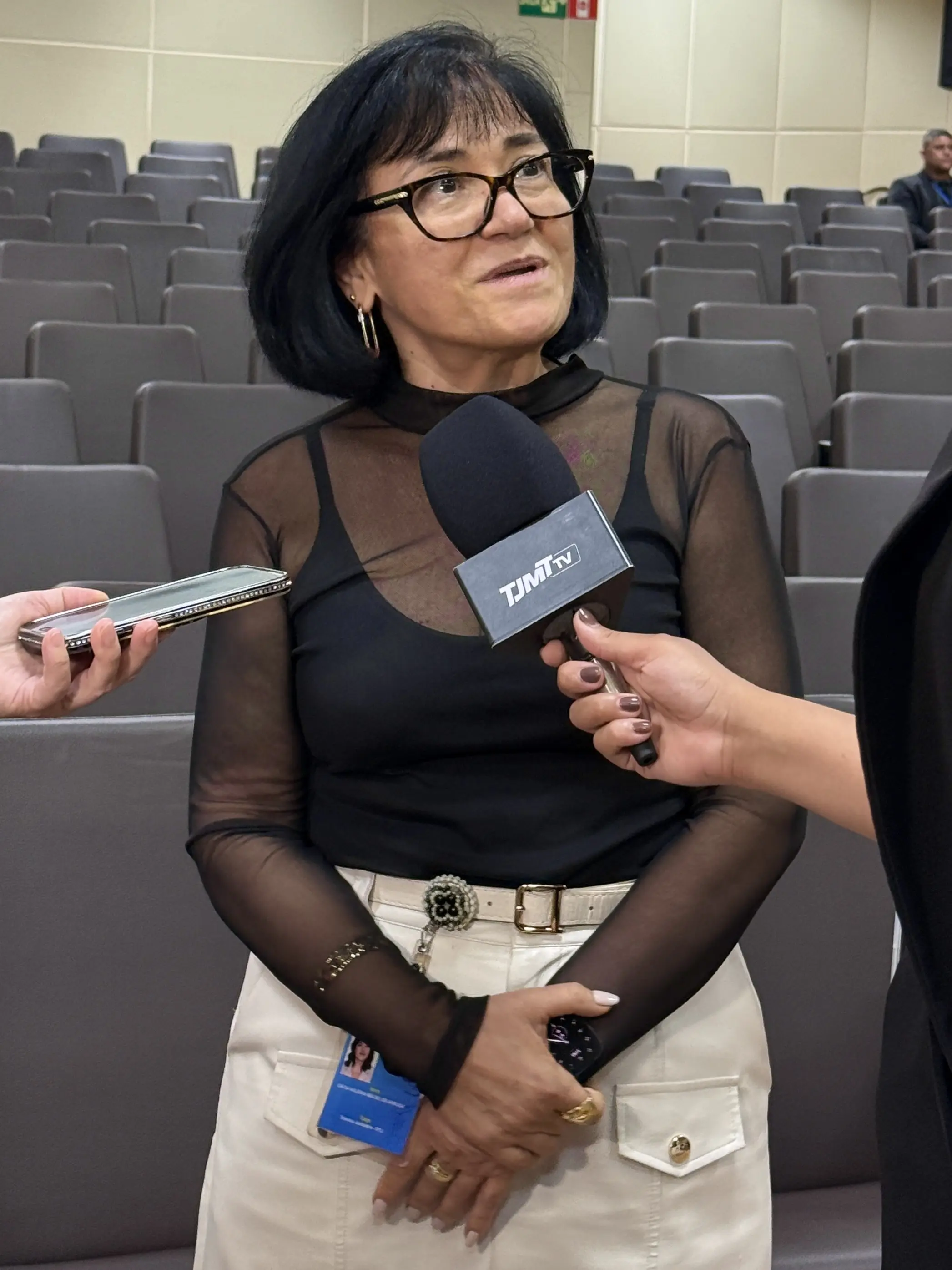 Mulher de cabelos pretos, óculos e blusa preta de tule, com crachá preso à saia clara, é entrevistada com microfone da TJMT TV em auditório. 