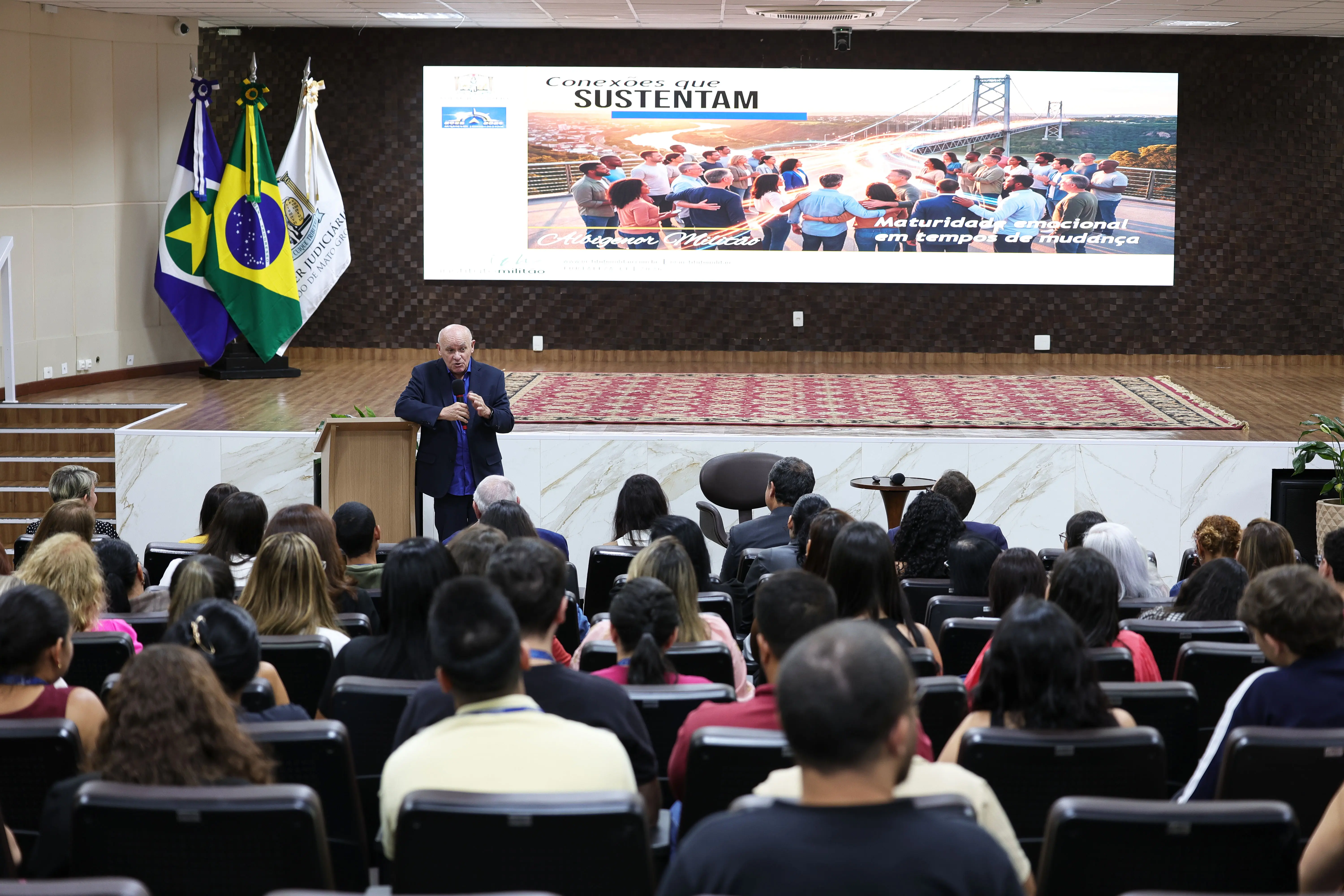 Homem calvo, de paletó azul-marinho e camisa azul, fala ao microfone em púlpito, diante da plateia e de telão com o tema “Conexões que sustentam”. 