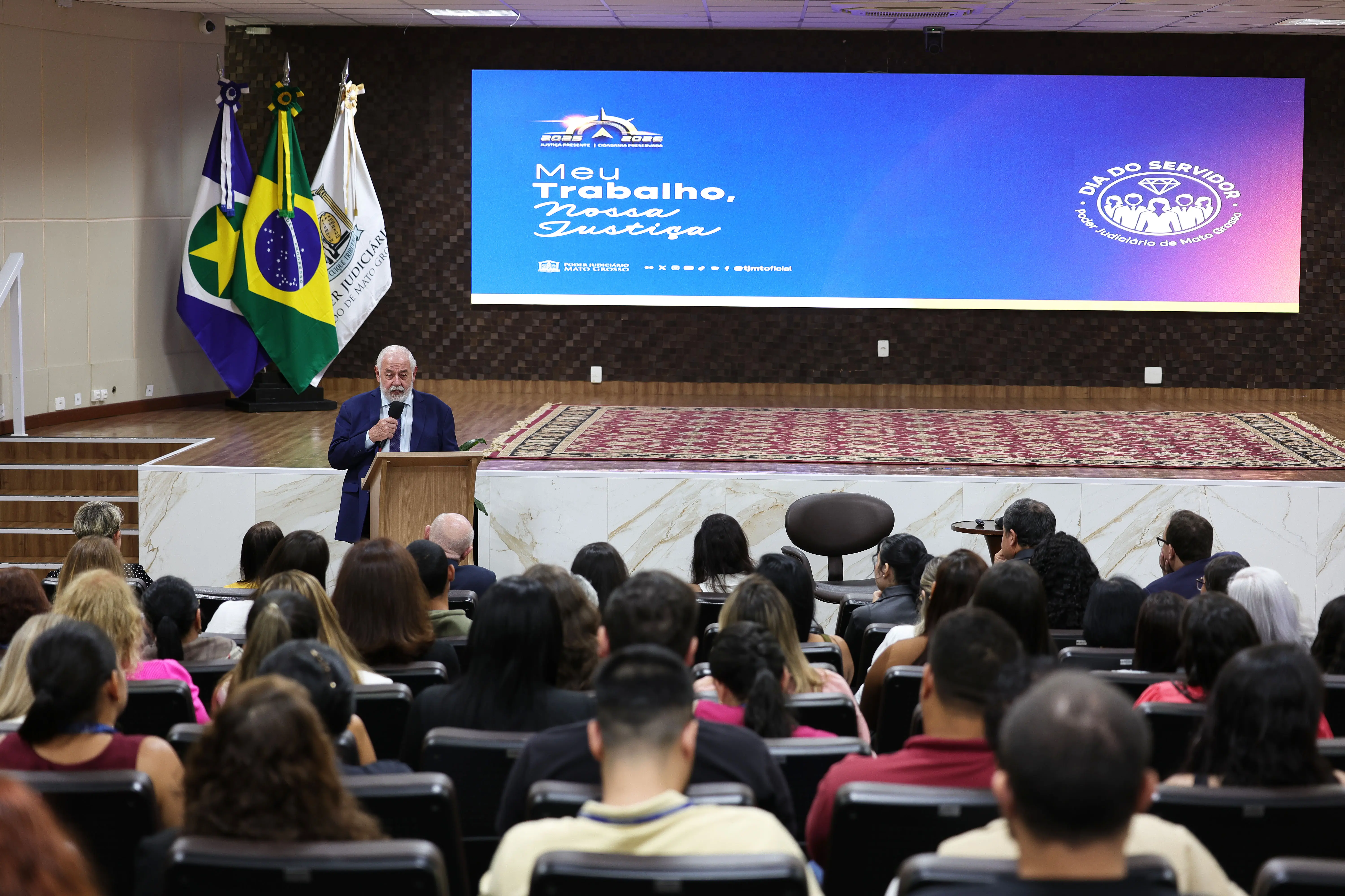 Homem de cabelos e barba brancos, de terno azul, discursa ao microfone em púlpito, diante da plateia e de telão com a arte do Dia do Servidor.