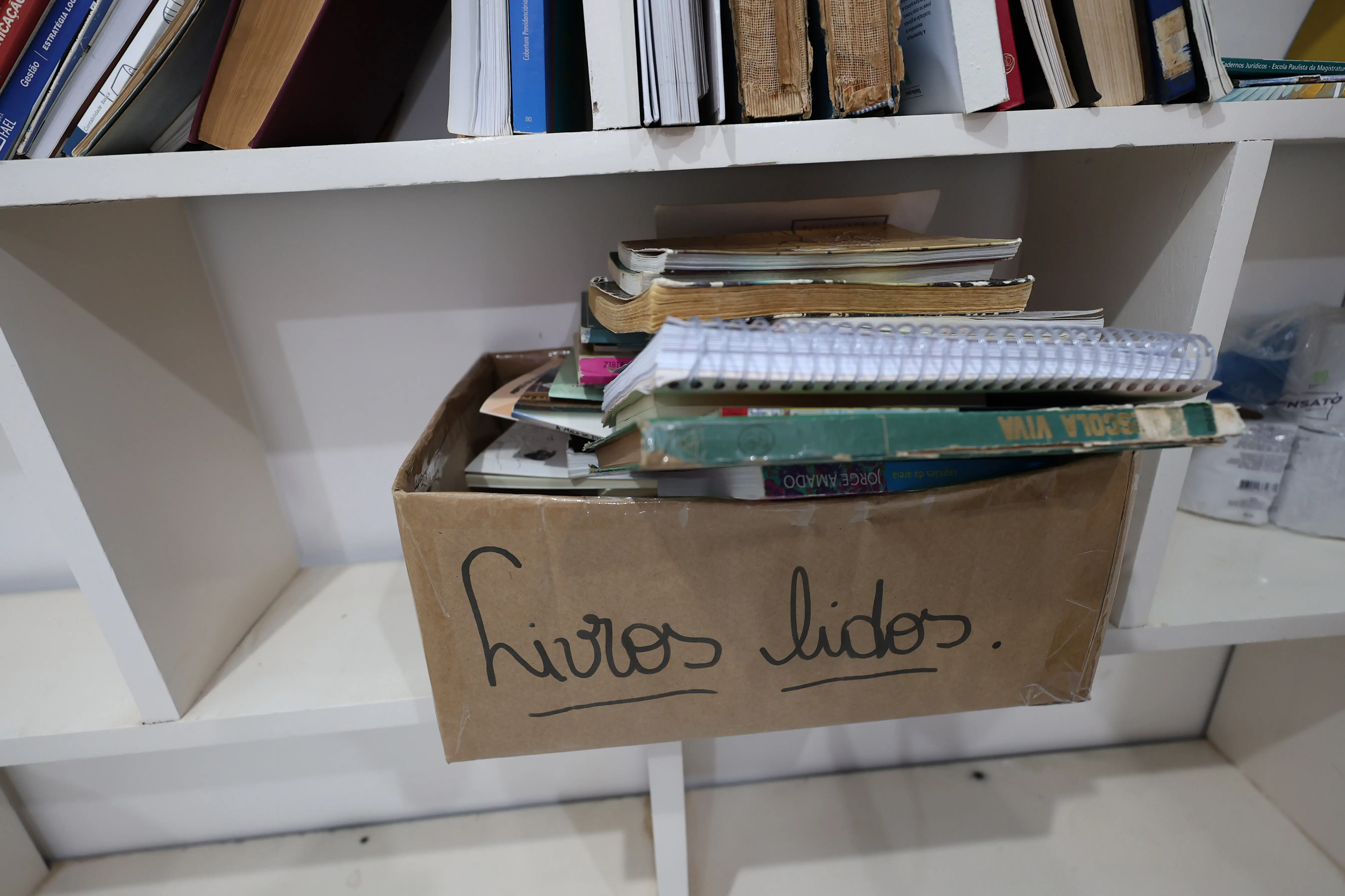 Caixa de papelão identificada como “livros lidos”, cheia de livros e cadernos, apoiada em uma estante branca com outros livros.