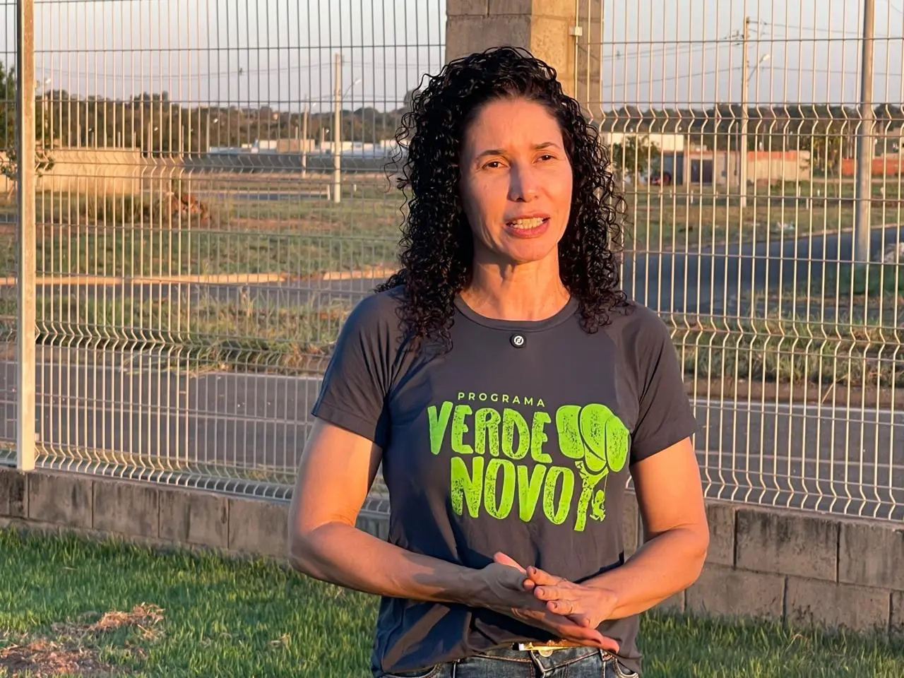 Mulher de cabelos cacheados escuros fala com as mãos unidas à frente, vestindo camiseta cinza do "Programa Verde Novo". Ao fundo, gramado e grade metálica sob iluminação solar.