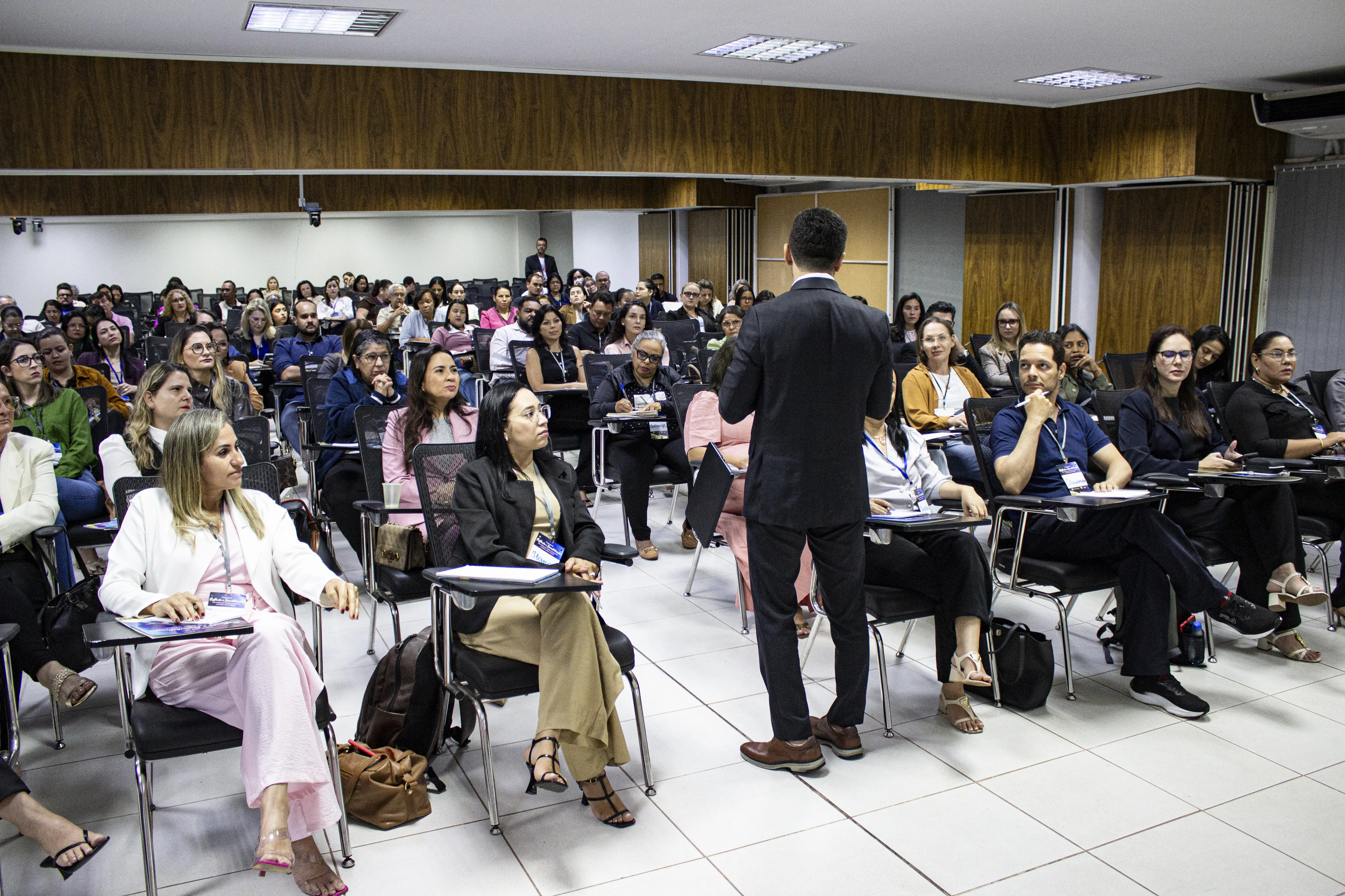Pessoa de costas fala para auditório com dezenas de participantes sentados em cadeiras escolares. O ambiente possui paredes amadeiradas e iluminação no teto.