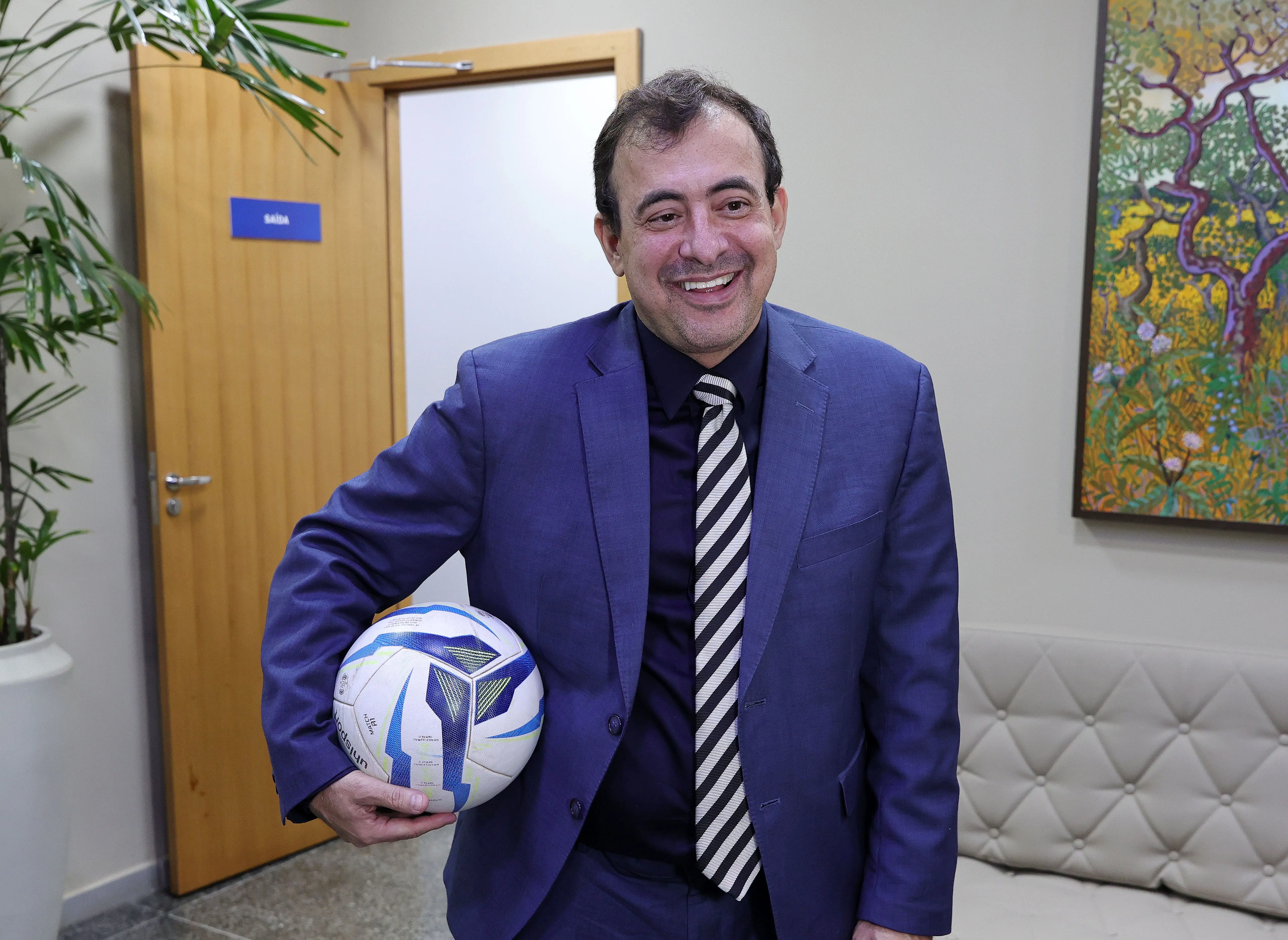 Foto horizontal que mostra o juiz  Jorge Alexandre Martins Ferreira da cintura pra cima, em pé, sorrindo e segurando uma bola de futebol. Ele é um homem branco, de cabelos castanhos, usando camisa azul marinho, terno azul e gravata listrada. 