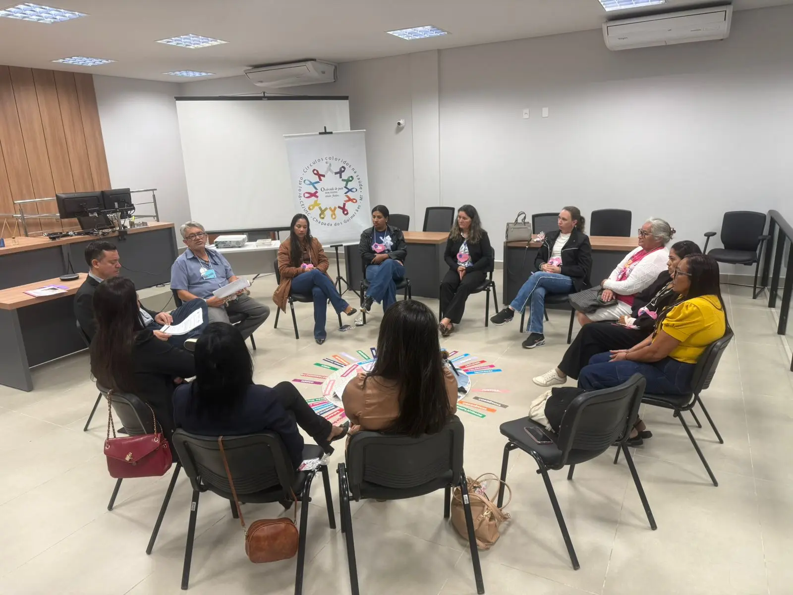 Foto horizontal em plano aberto que mostra um grupo de 12 pessoas sentadas em círculo em uma sala no fórum. Ao fundo há um banner com a logomarca do projeto Círculos Coloridos na Saúde.