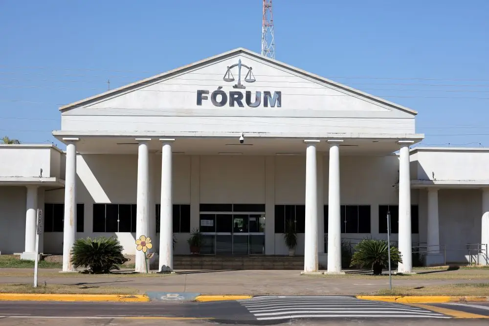 Foto horizontal em plano aberto que mostra a fachada do Fórum de Sinop em uma dia de céu azul ensolarado. A fachada tem estilo neoclássico, com seis colunas dóricas que sustentam a fachada triangular onde está o letreiro escrito Fórum e a balança da Justiça.