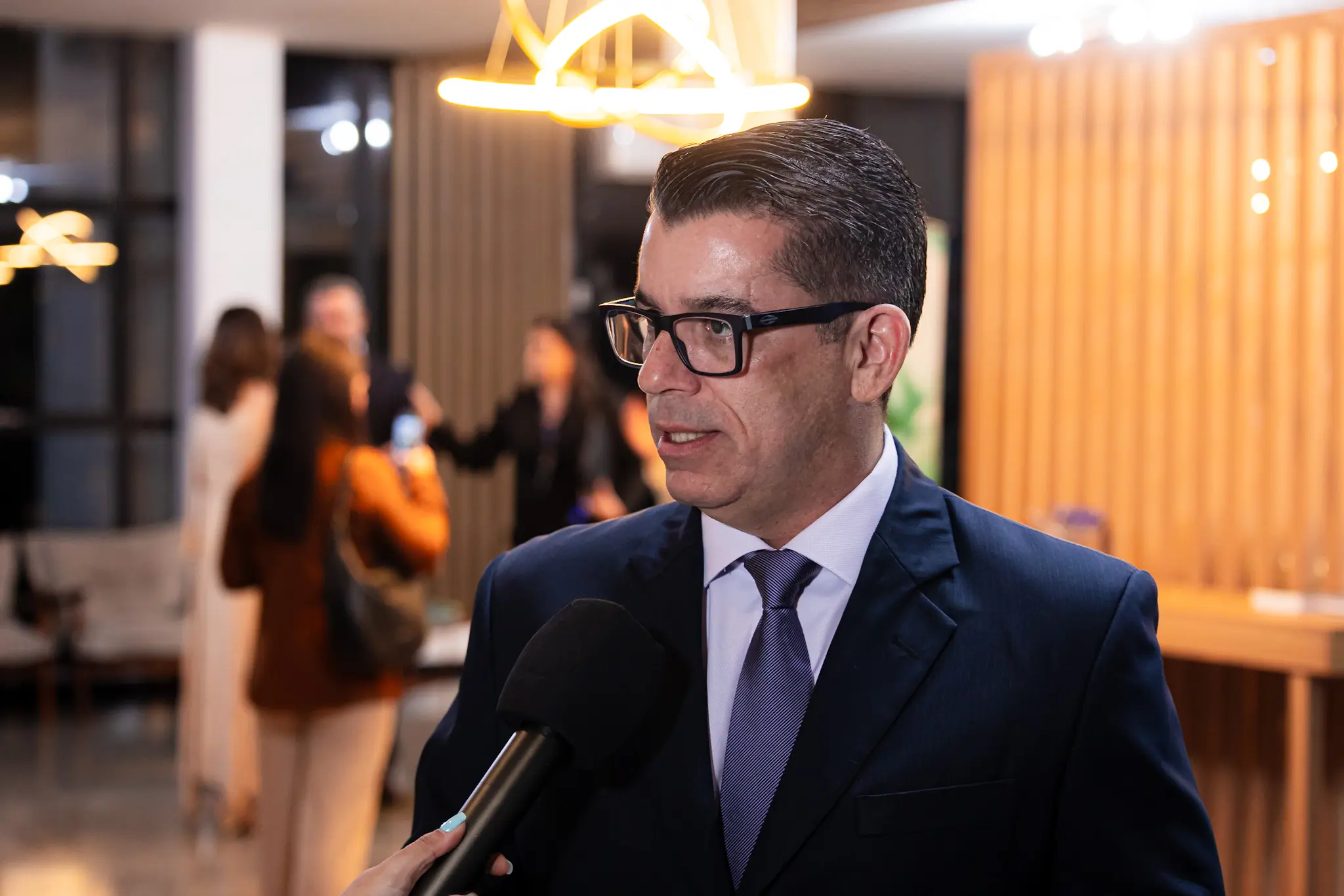Homem de óculos de armação escura e cabelos curtos concede entrevista. Ele veste terno escuro, camisa clara e gravata em tom roxo.
