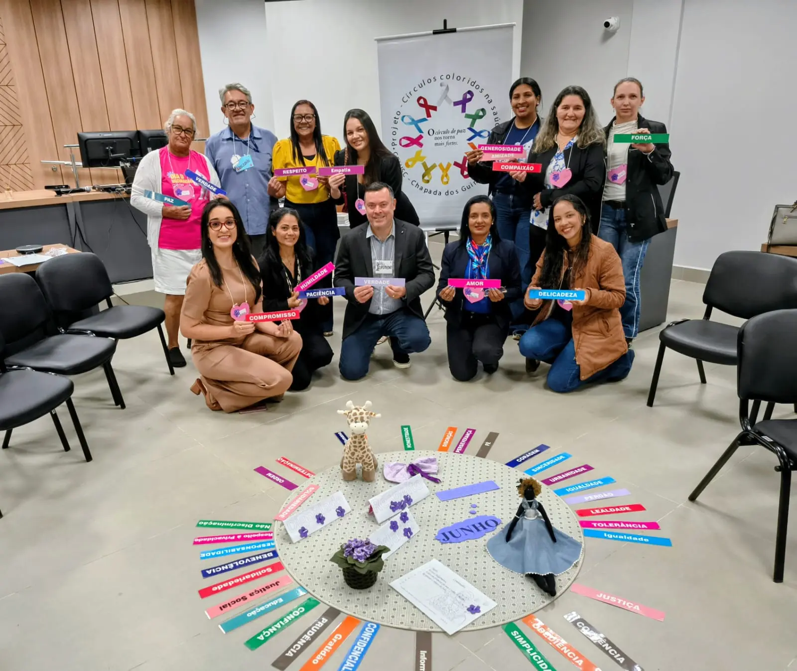 Foto horizontal em plano aberto que mostra um grupo de 12 pessoas posando sorrindo para a foto, uma parte em pé e outra agachada, segurando fitas de papel coloridos com palavras variadas que simbolizam diversas virtudes humanas. No chão, há outros papeis coloridos e outros objetos sobre um tapete.