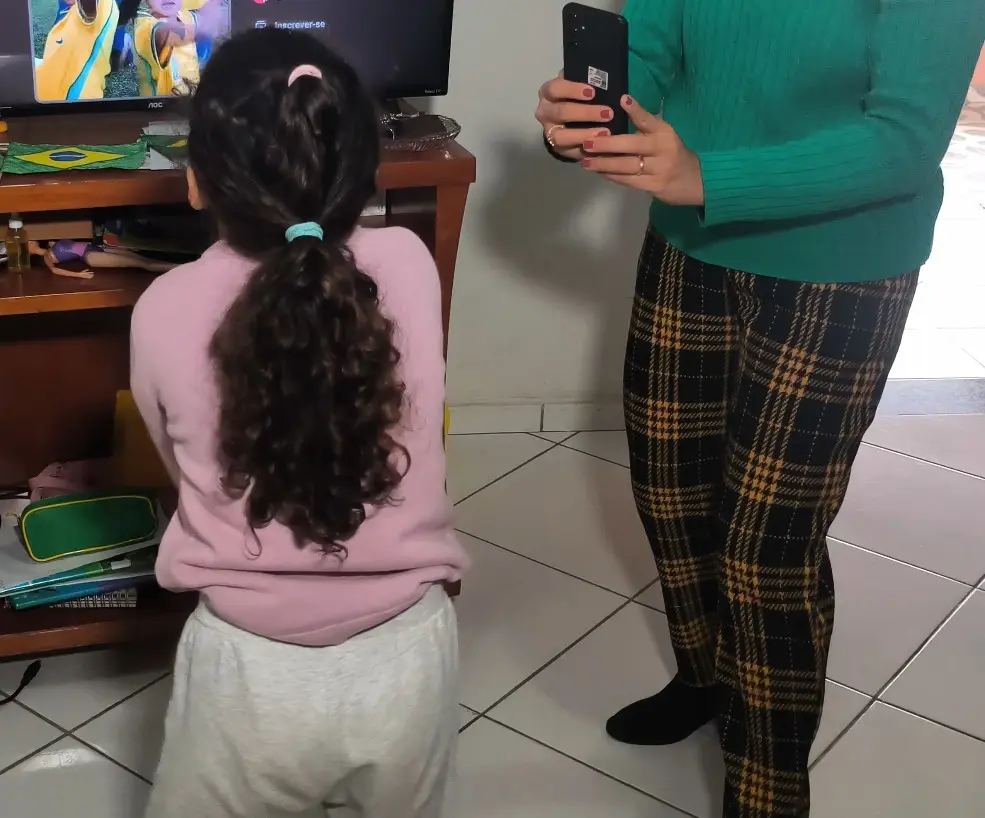 Foto horizontal que mostra uma menina sendo filmada dançando na frente da televisão. Quem filma com o celular é uma mulher usando blusa de frio verde e calça xadrez preta e amarela. A menina aparece de costas, com cabelos longos, castanhos e cacheados presos, usando calça cinza e moletom rosa.
