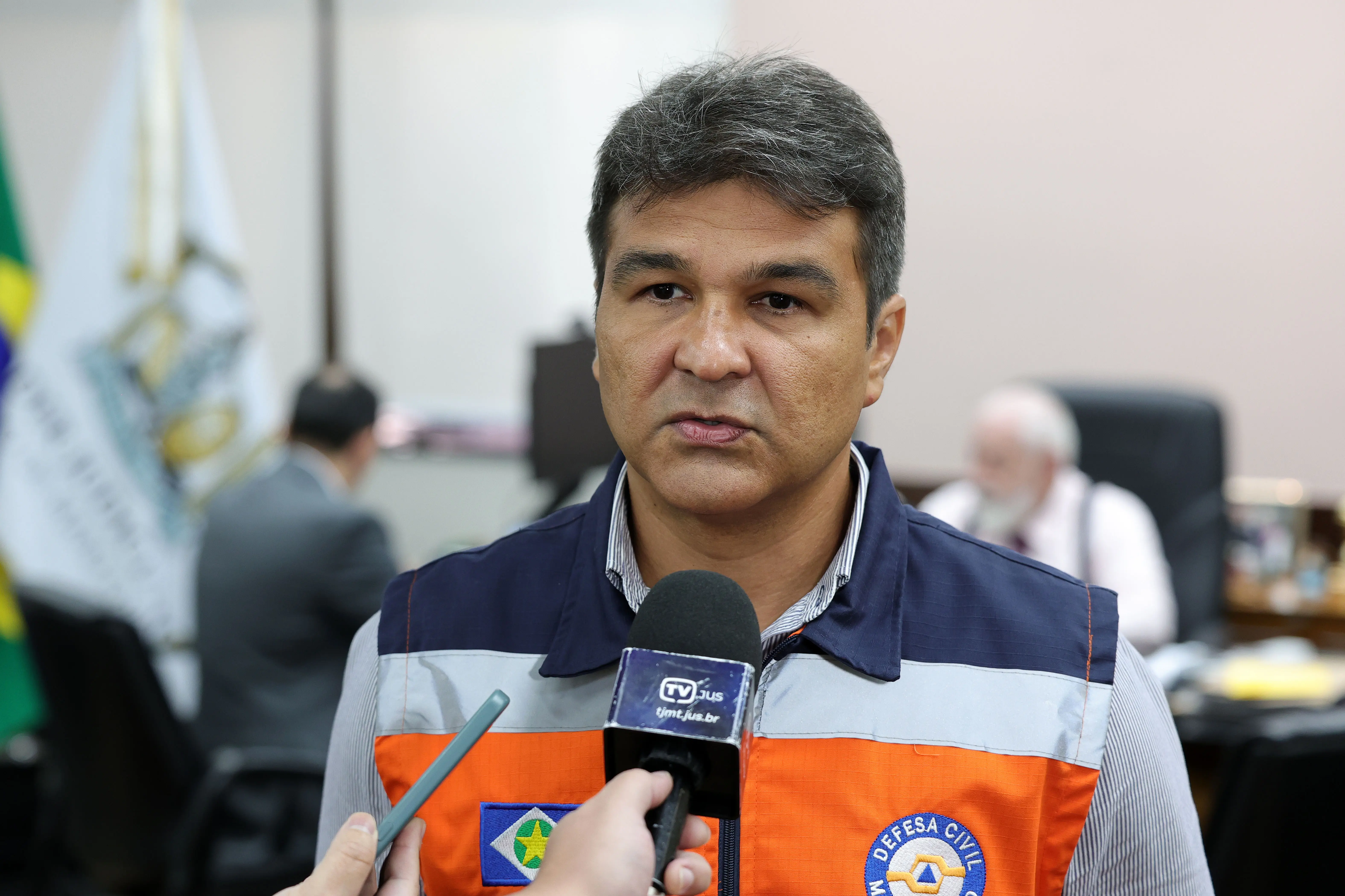 Homem de cabelos curtos grisalhos veste colete da Defesa Civil de Mato Grosso e concede entrevista. Um microfone da TV JUS e um celular são apontados para ele. Fundo de escritório desfocado.