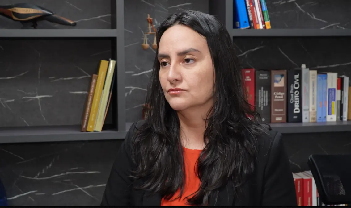Mulher de cabelos longos e lisos pretos, veste blazer preto sobre blusa laranja, olha para o lado. Ao fundo, estante escura com livros jurídicos, entre eles Código Penal e Direito Civil.