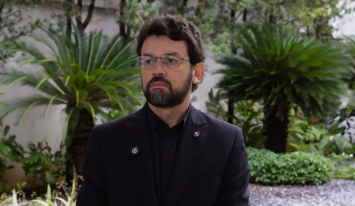 Foto horizontal que mostra o juiz Tiago Abreu, do busto pra cima. Ele é um homem branco, de cabelo, barba e olhos castanho escuros, usando óculos de grau, camisa e terno pretos. Ao fundo, várias plantas verdes no jardim do tribunal. 