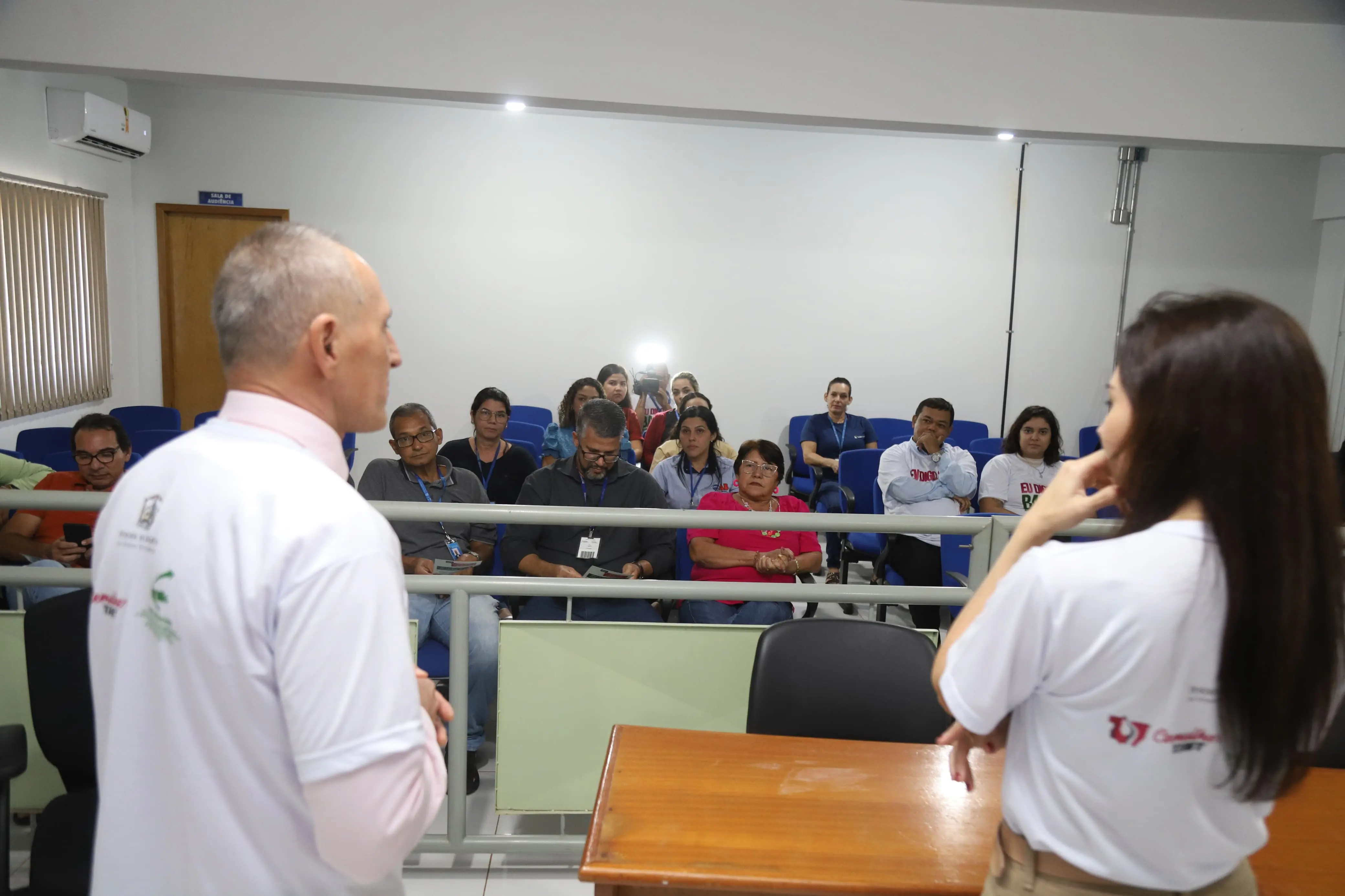 Homem e mulher falam a um grupo sentado em auditório. Os participantes observam com atenção. Alguns usam camisetas da campanha “Eu Digo Basta!”. Ao fundo, uma pessoa fotografa com flash. Ambiente iluminado, com cadeiras azuis e parede branca.