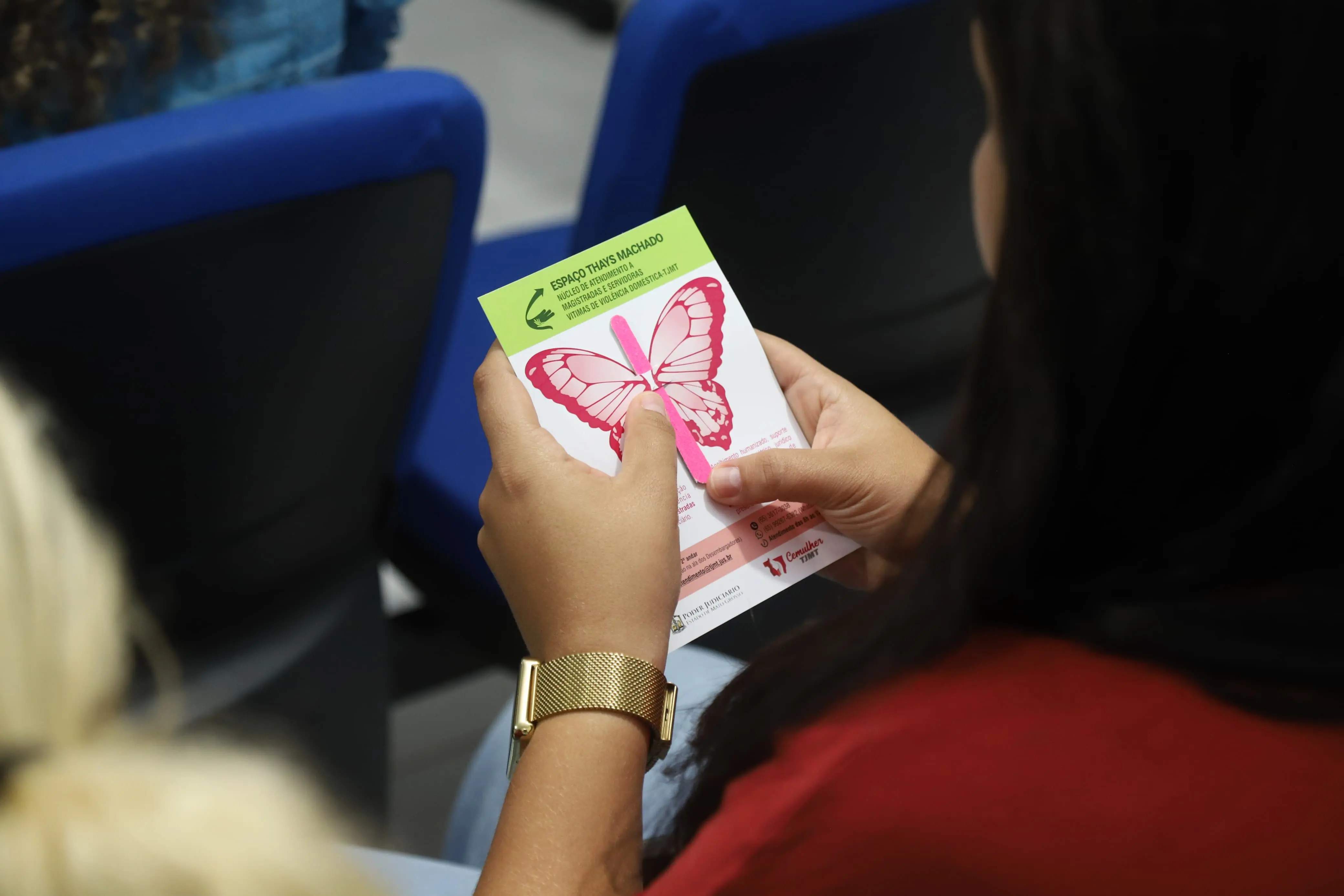 Mulher segura um folder com desenho de borboleta rosa e informações sobre o Espaço Thaís Machado, que acolhe vítimas de violência doméstica. A imagem destaca o material informativo em meio à plateia, com cadeiras azuis ao fundo.