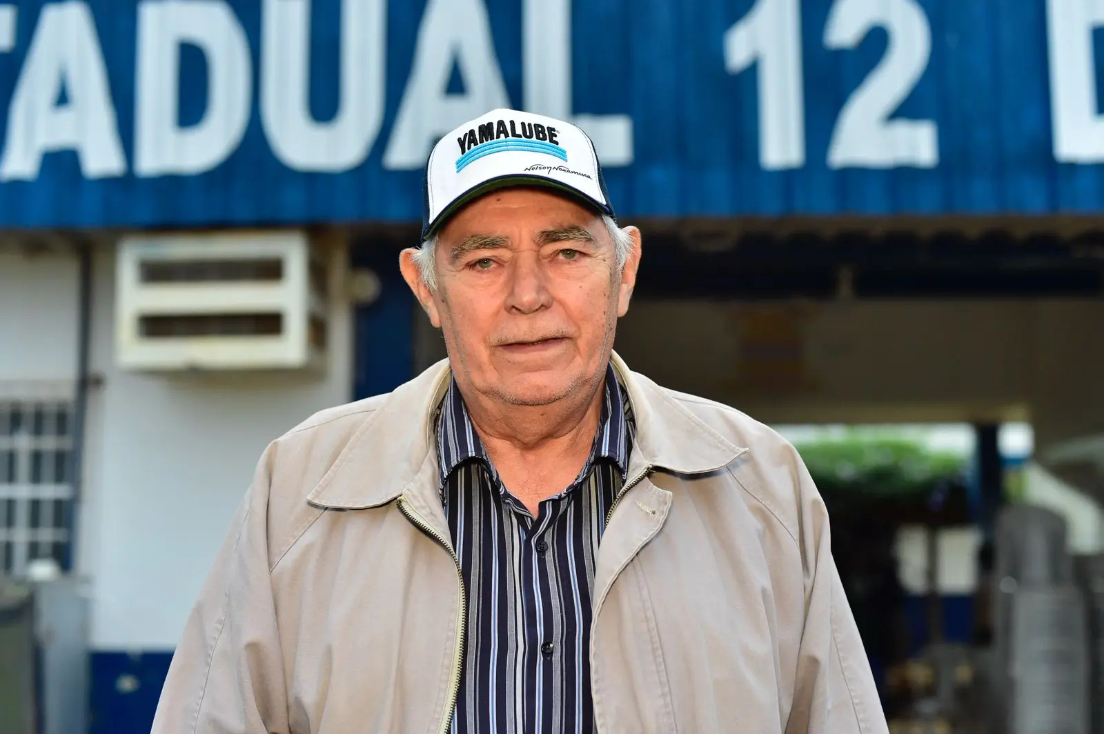 Homem idoso de boné e jaqueta bege posa em frente à fachada da Escola Estadual 12 de Outubro. Parte da parede azul com letras brancas da escola aparece ao fundo.