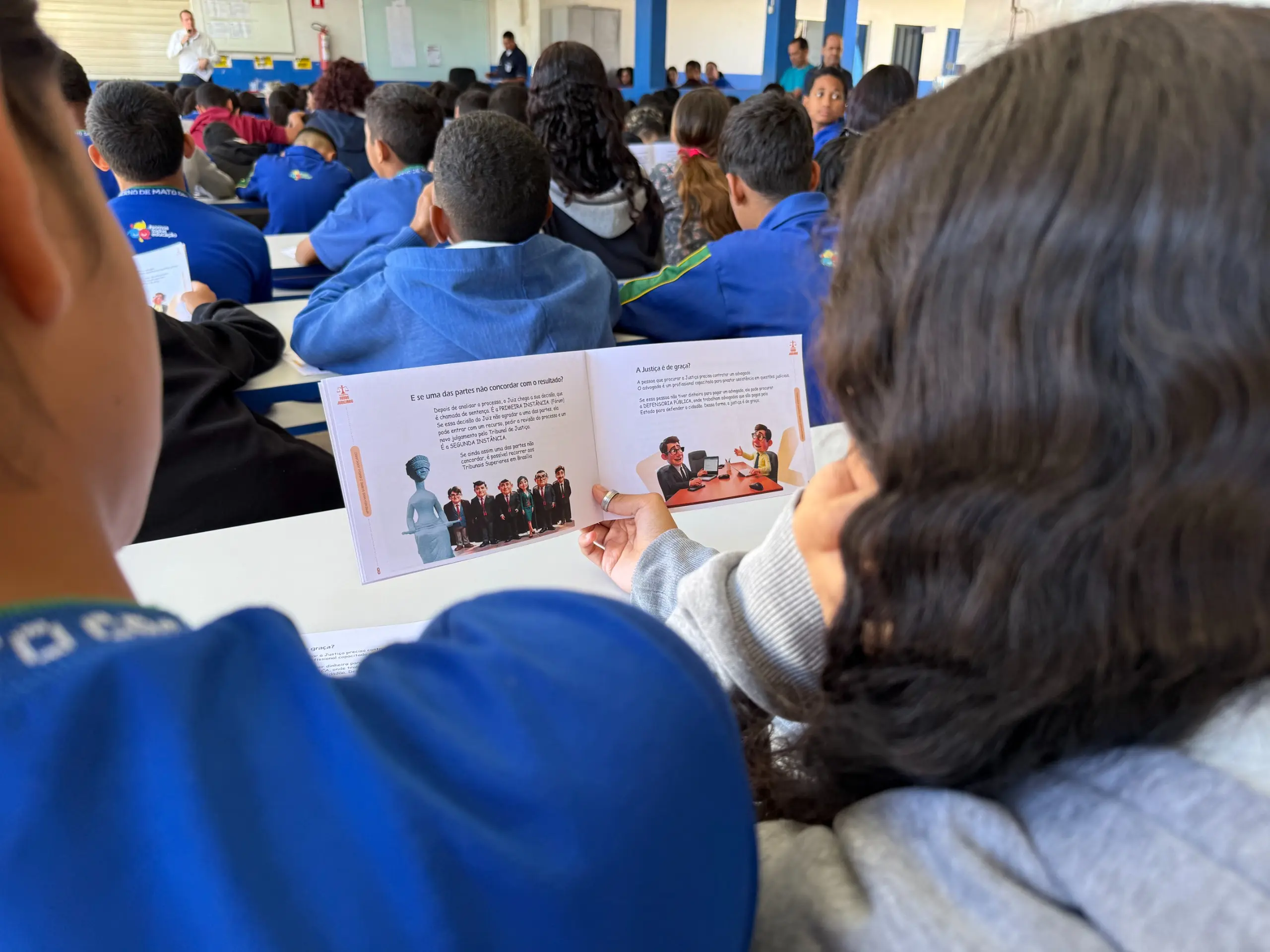 A câmera foca em um folheto informativo aberto sobre uma mesa, que é segurado por duas mãos. O folheto mostra duas ilustrações: uma de um grupo de pessoas em frente a um tribunal e outra de um juiz em seu gabinete. 