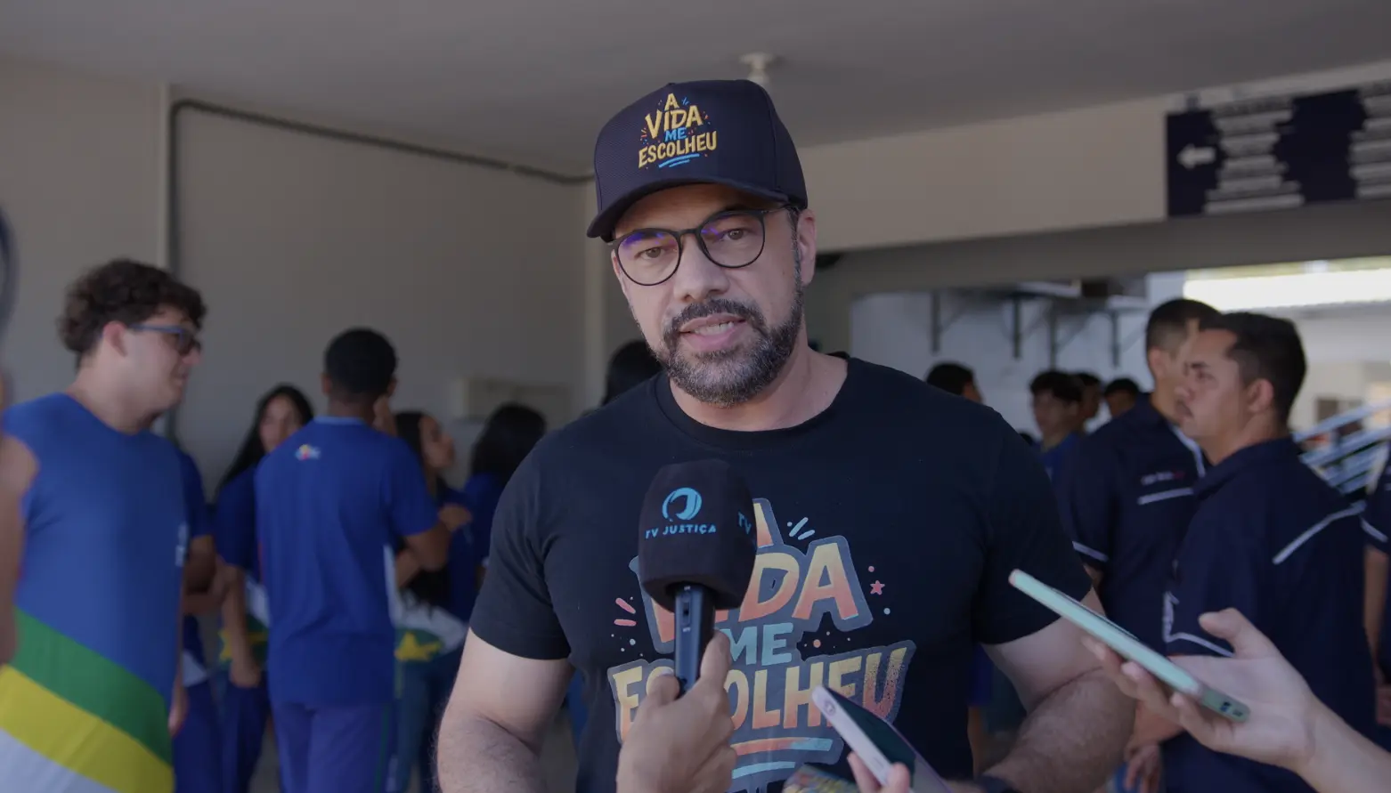 Psicólogo Afro Stefanini concede entrevista à TV Justiça. ele é um homem pardo, de barba, usando camiseta e boné pretos com a logomarca do projeto A vida me Escolheu. Ao fundo, alunos no pátio de uma escola estadual.