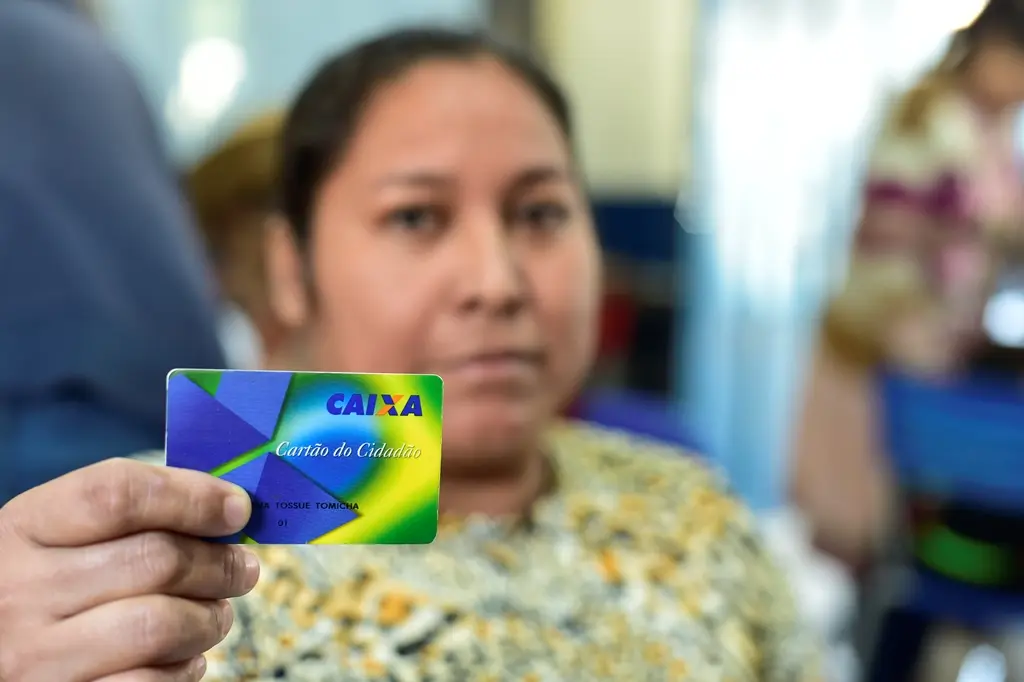 Mulher mostra o cartão do cidadão, da Caixa Econômica Federal. Ela está desfocada e o cartão em primeiro plano.