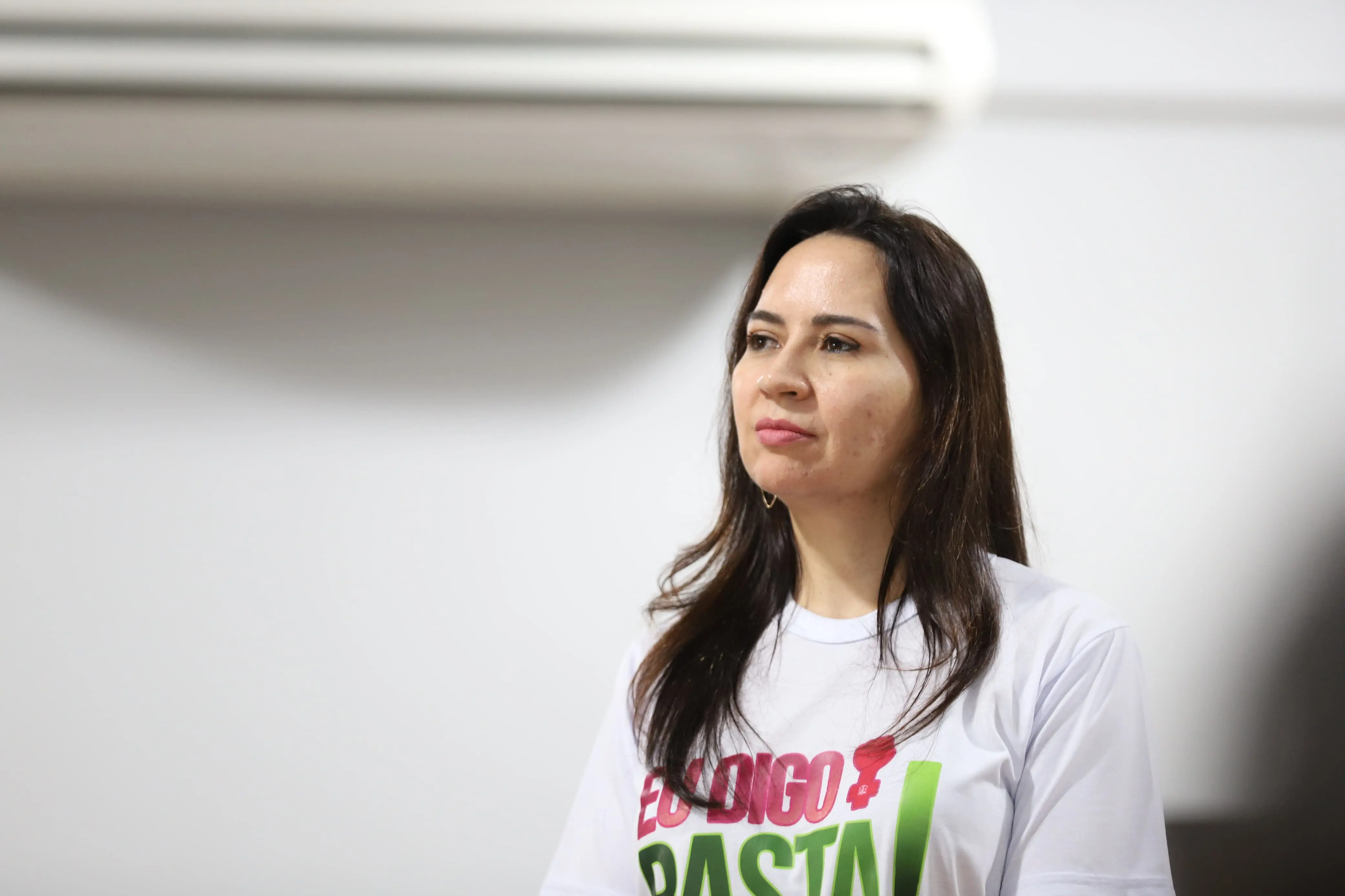 Mulher de cabelos castanhos escuros e lisos veste camiseta branca da campanha “Eu Digo Basta!”. Ela está em pé, com expressão séria e atenta, diante de uma parede clara com ar-condicionado ao fundo. A imagem transmite foco e firmeza.