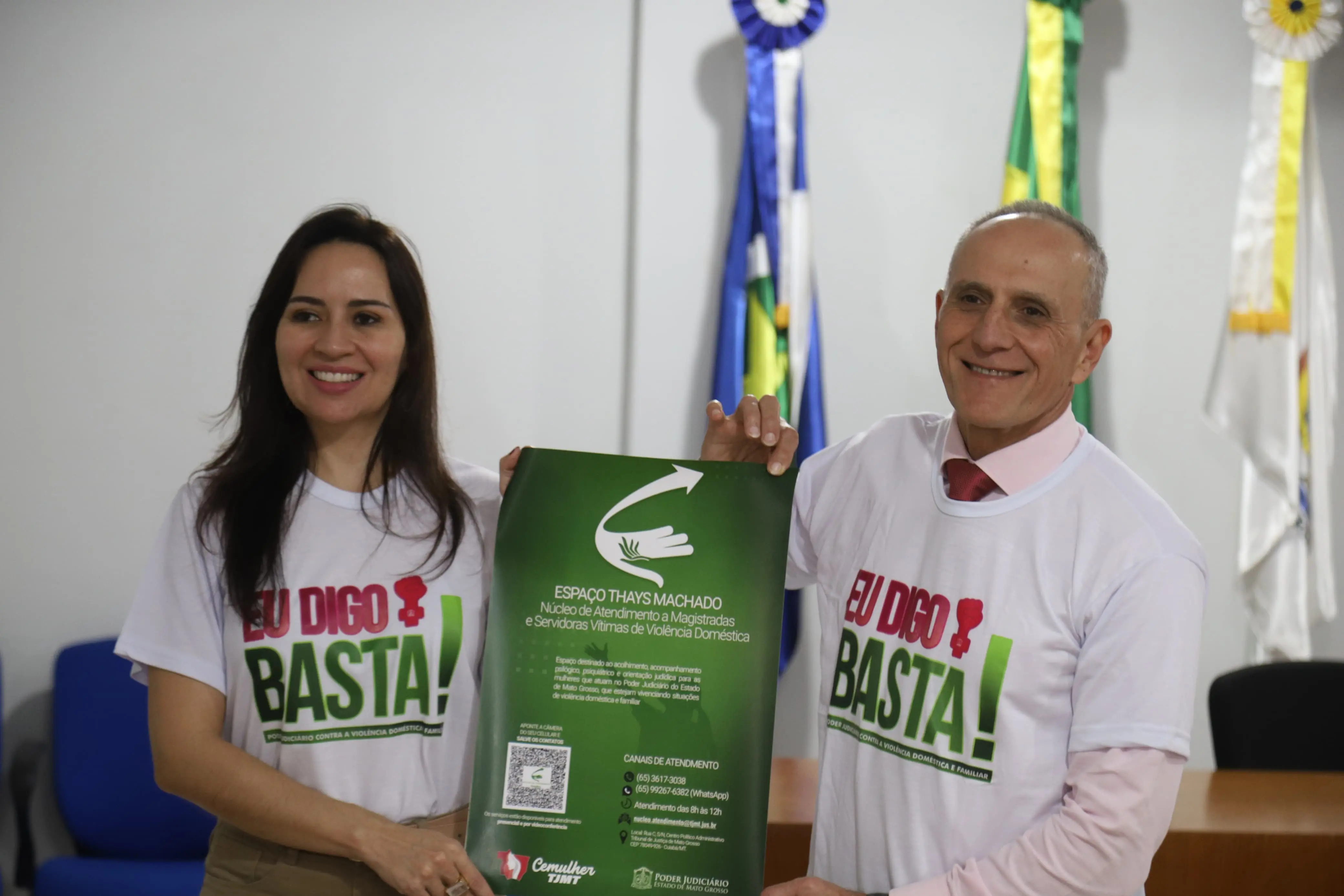Mulher e homem sorriem enquanto seguram cartaz verde do Espaço Thays Machado. Ambos vestem camisetas da campanha “Eu Digo Basta!”. Ao fundo, aparecem bandeiras institucionais e cadeiras azuis, em ambiente de evento oficial.