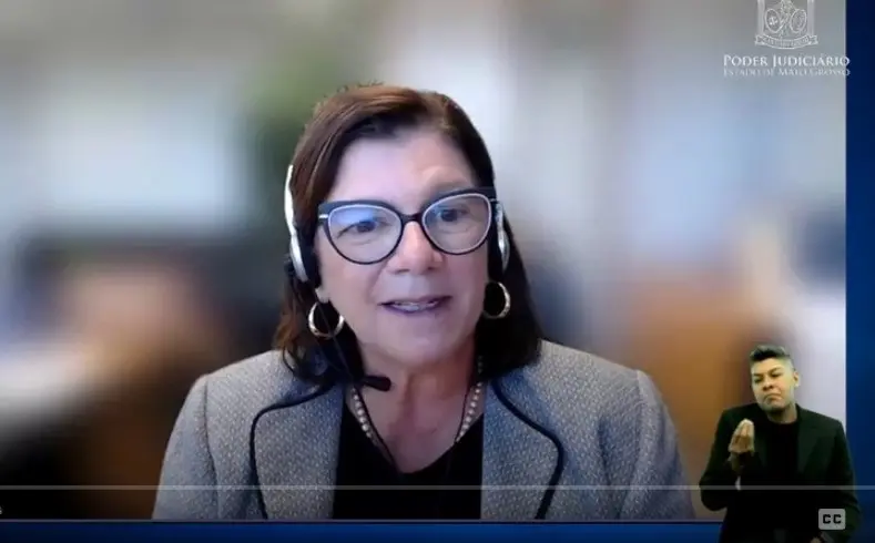 Uma mulher, palestrante, usa fones de ouvido e óculos, falando em um webinário. No canto inferior direito, um intérprete de Libras. O logo do "Poder Judiciário Estado de Mato Grosso" está no canto superior direito.