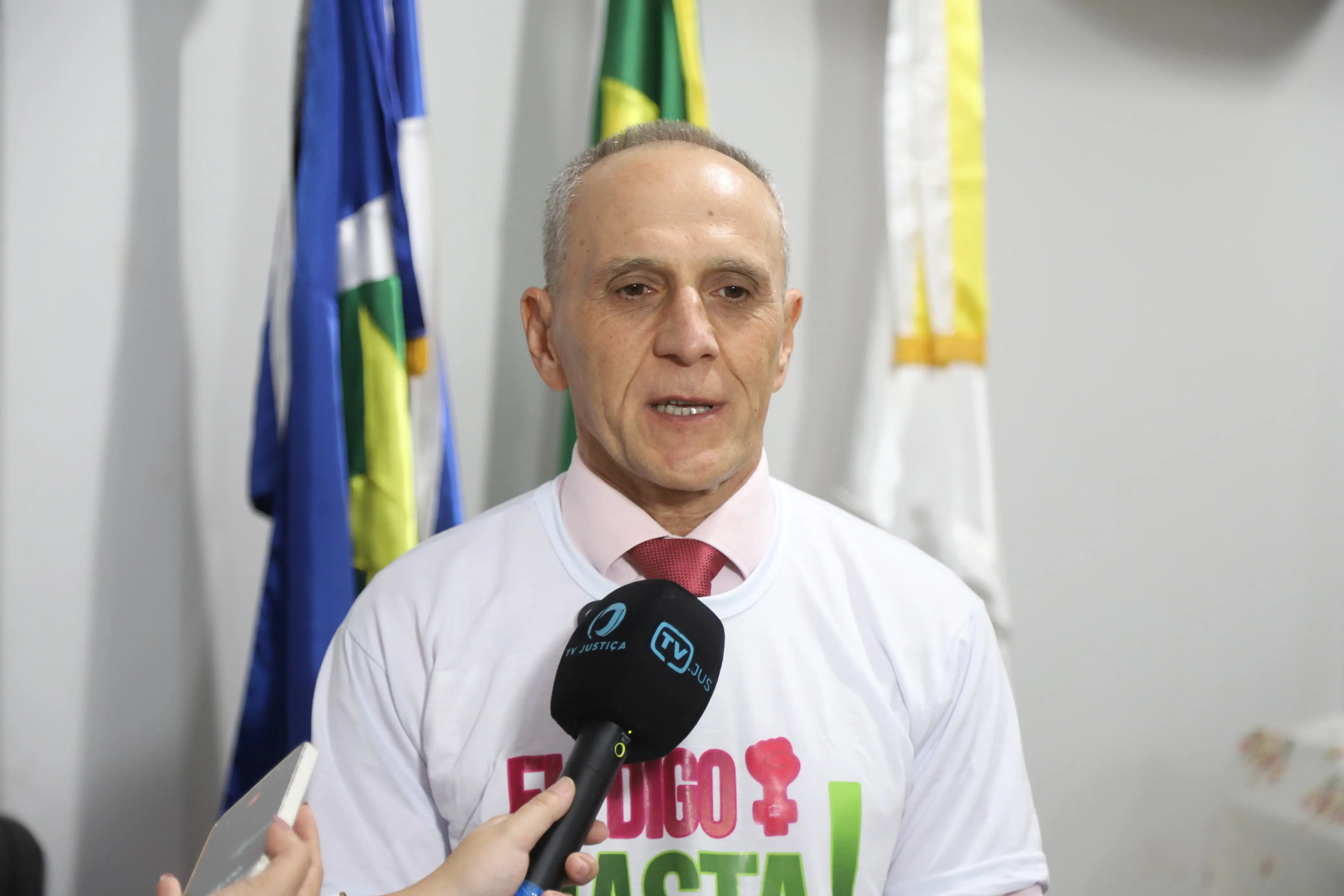 Homem de terno e gravata vermelha, vestindo camiseta da campanha “Eu Digo Basta!”, concede entrevista à imprensa. Ele fala diante de microfone com logo da TV Justiça. Ao fundo, aparecem bandeiras institucionais e parede clara.