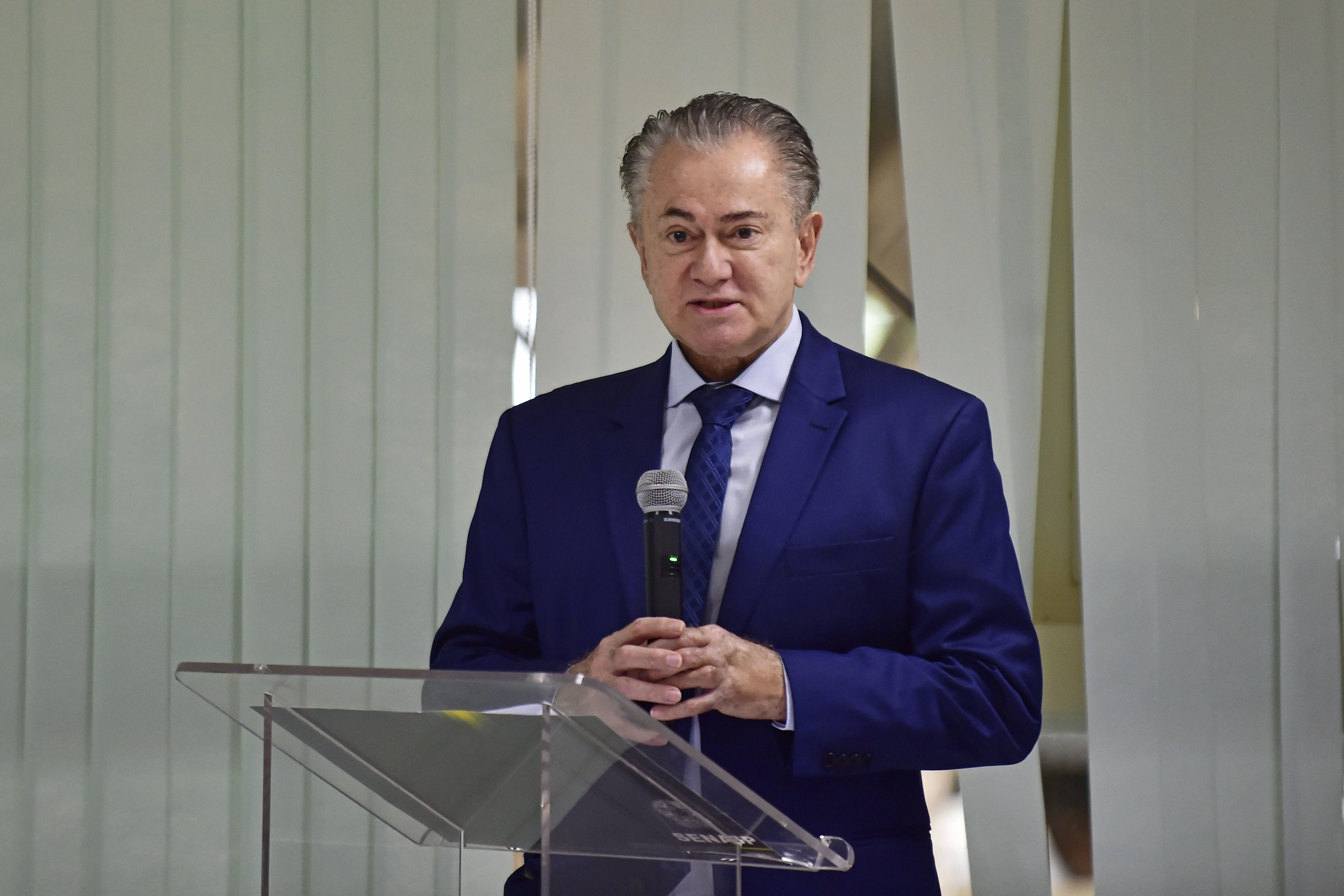 Desembargador Orlando Perri fala ao microfone, em um púlpito de acrílico transparente. Ele é um senhor branco, de cabelos grisalhos, usando camisa azul clara, tenho e gravata azul-marinho. 