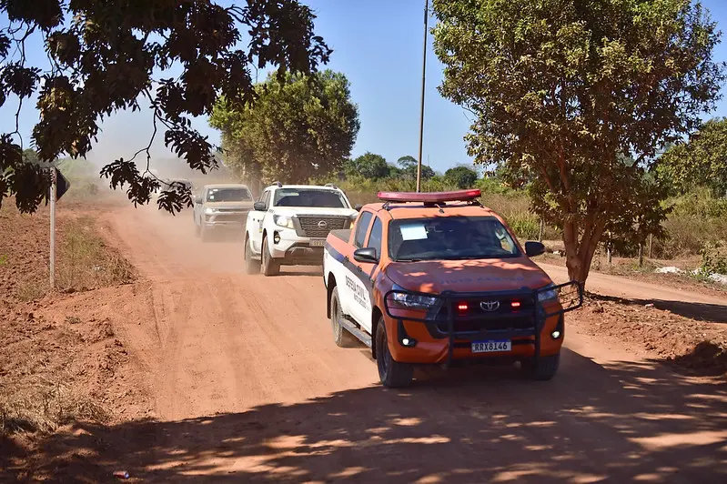 Comboio liderado pela caminhonete da Defesa Civil trafega por uma estrada de chão, cheia de poeira.