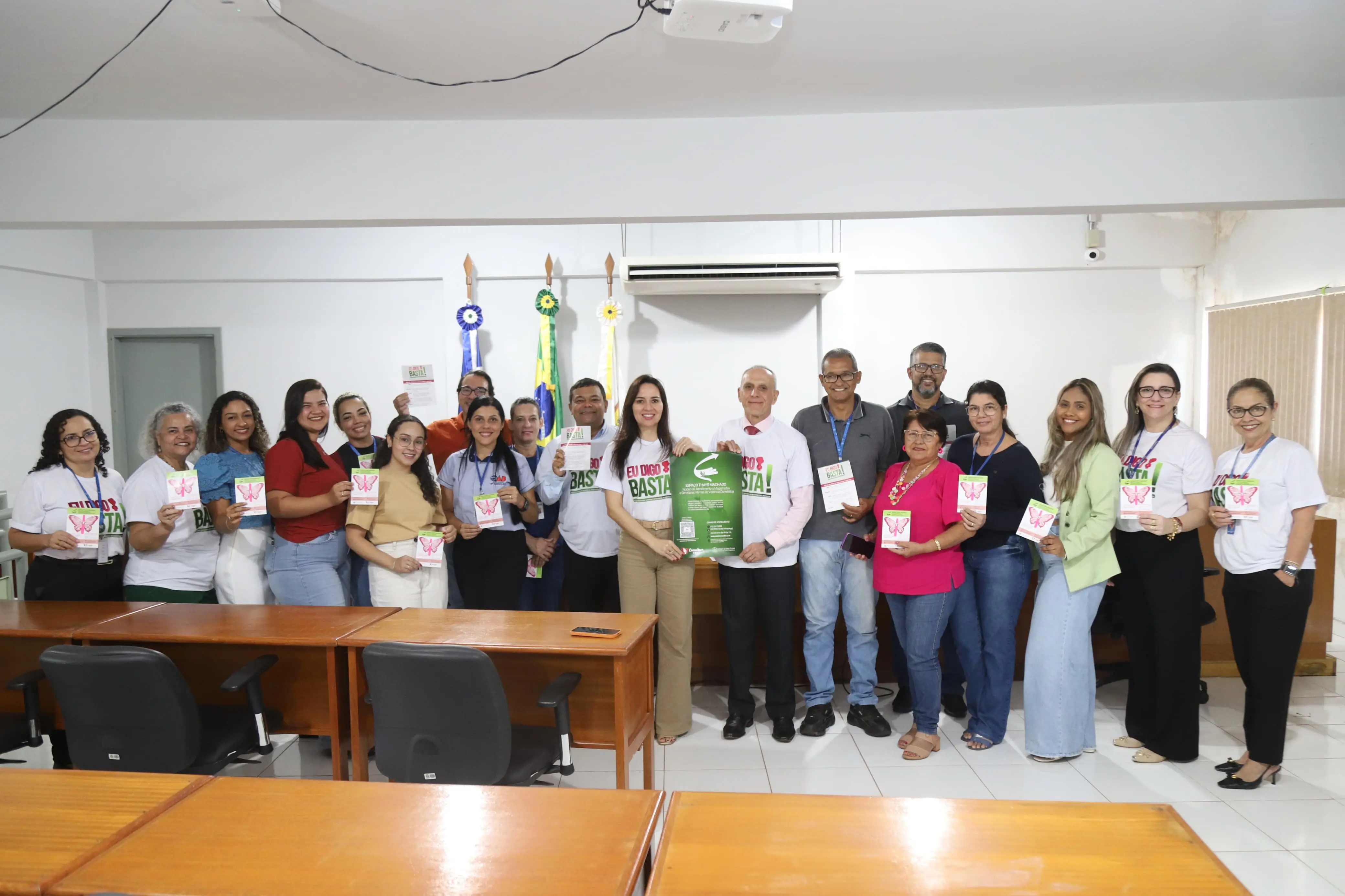 Grupo de 21 pessoas posa sorridente em sala de audiência, segurando folders com borboleta rosa, símbolo da campanha “Eu Digo Basta!”. Ao centro, duas pessoas exibem cartaz verde. Ao fundo, bandeiras oficiais e ar-condicionado fixado na parede.
