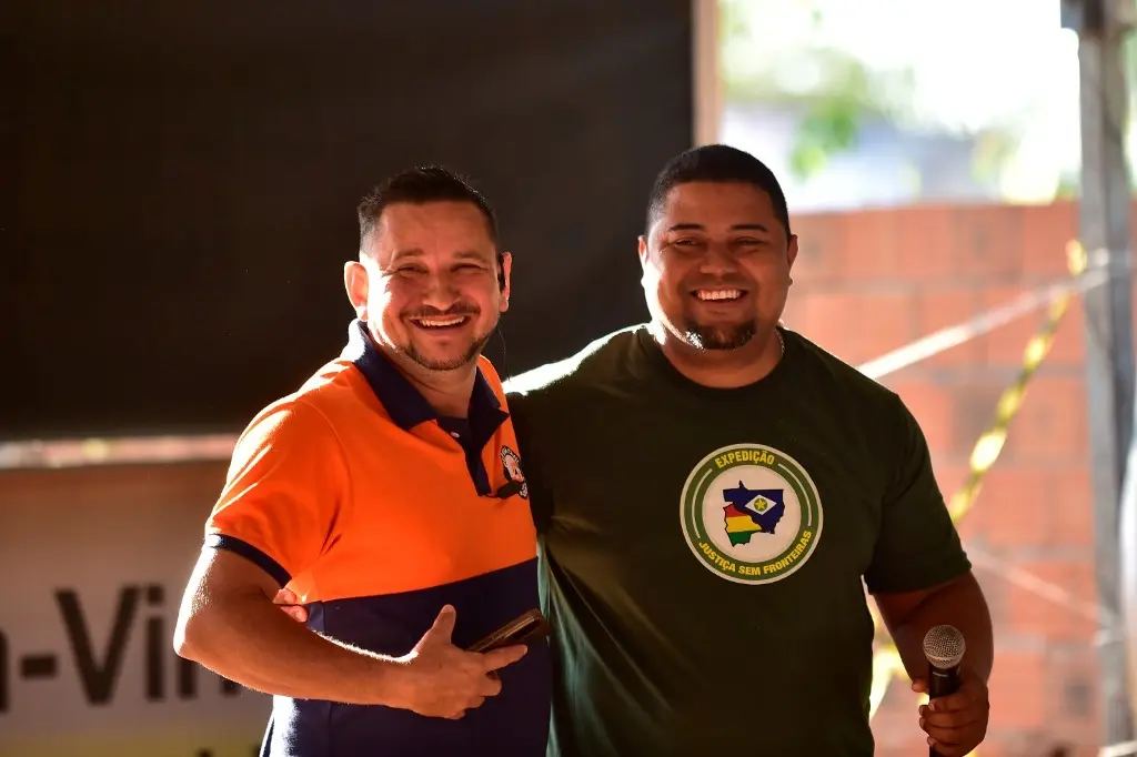 Dois voluntários da Justiça Sem Fronteiras posam sorrindo para a foto. Um deles é um homem branco, usando camiseta azul e laranjada da Defesa Civil. O outro é um homem negro, usando camiseta verde da Justiça Sem Fronteiras.