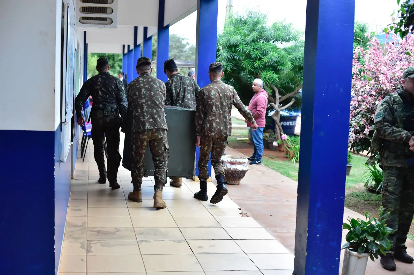 Militares fardados caminham por corredor de escola carregando equipamento pesado. Ao lado, homem observa e vegetação com flores decora o ambiente externo