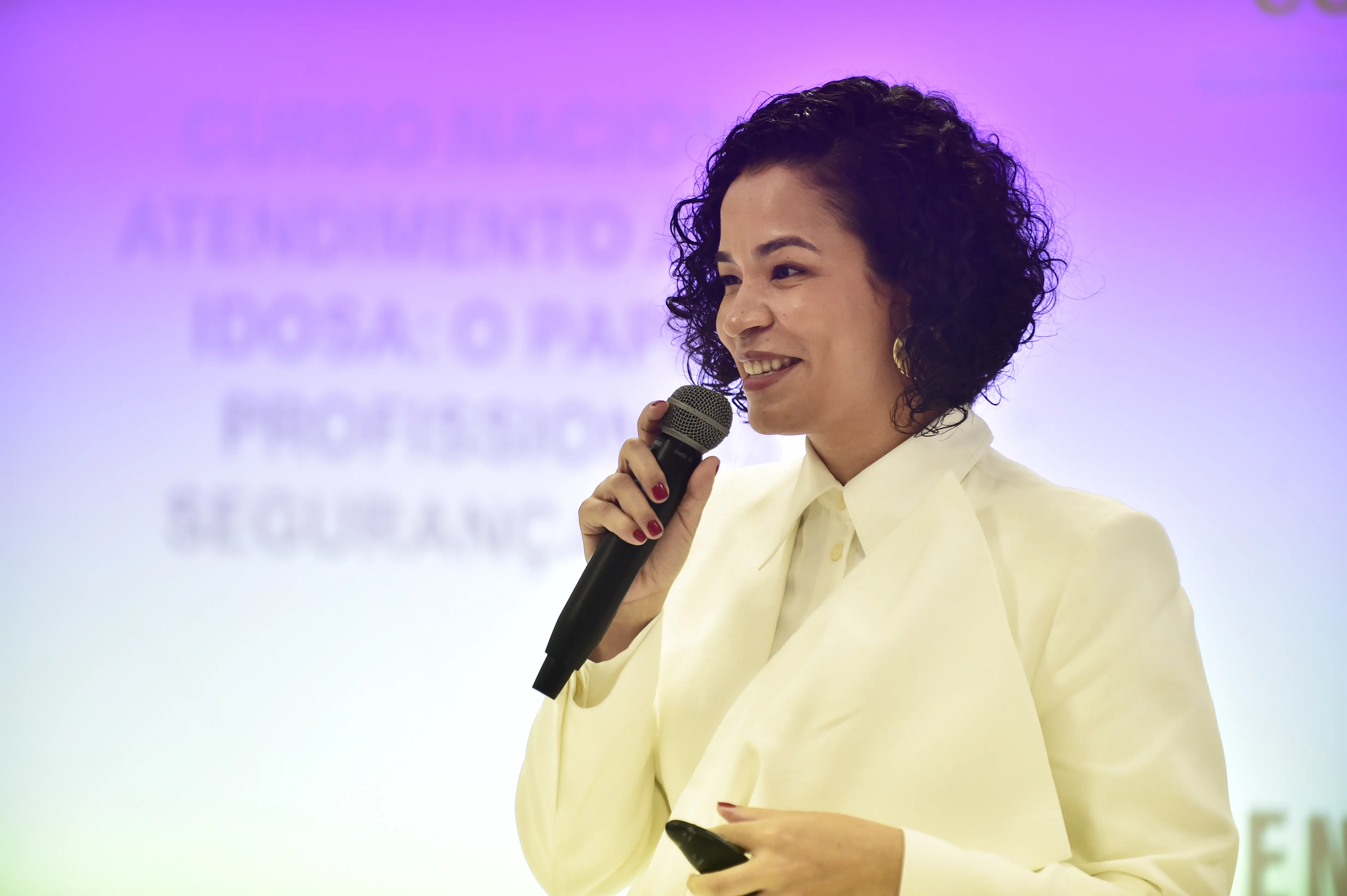 Foto que mostra a professora Rose Ferreira durante a aula, falando ao microfone e sorrindo. Ela é uma mulher parda, de olhos castanhos, cabelo preto, cacheado com corte long bob. Ela usa blazer off-white e brincos de argola dourados. 