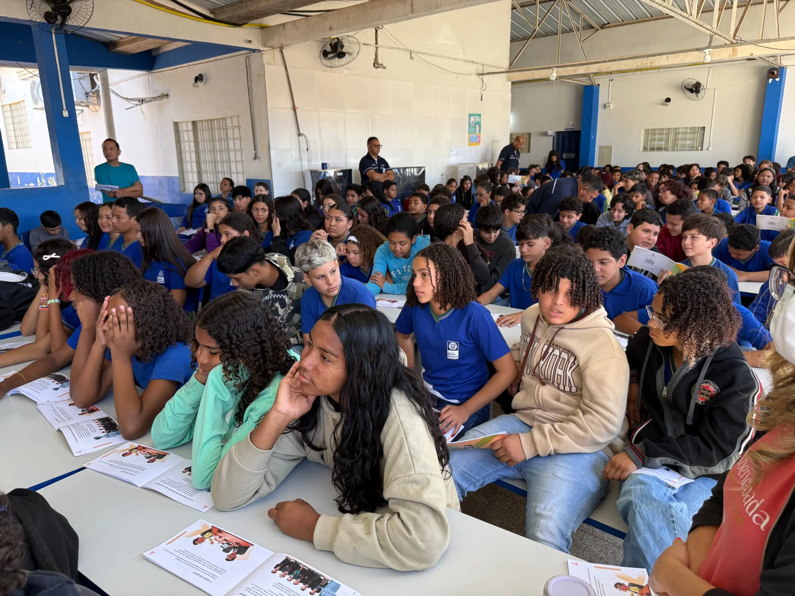  Vários estudantes, a maioria vestindo camisetas azuis, estão sentados em mesas, participando de uma palestra. Alguns olham para frente, para a pessoa que está falando, enquanto outros conversam ou olham para baixo, onde estão folhetos sobre a mesa. A cena é de uma atividade educativa em grupo.