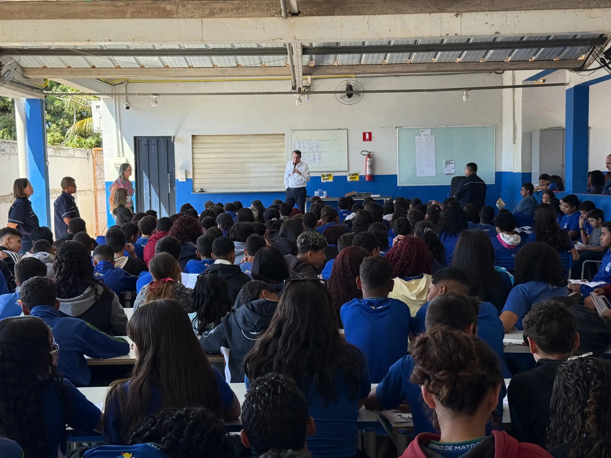 Esta imagem mostra uma visão de trás de uma grande plateia de estudantes. Eles estão sentados em fileiras, com suas costas voltadas para a câmera, ouvindo uma apresentação. A sala é ampla, com um teto de estrutura metálica e paredes azuis, e ao fundo, um grupo de adultos supervisiona a atividade.