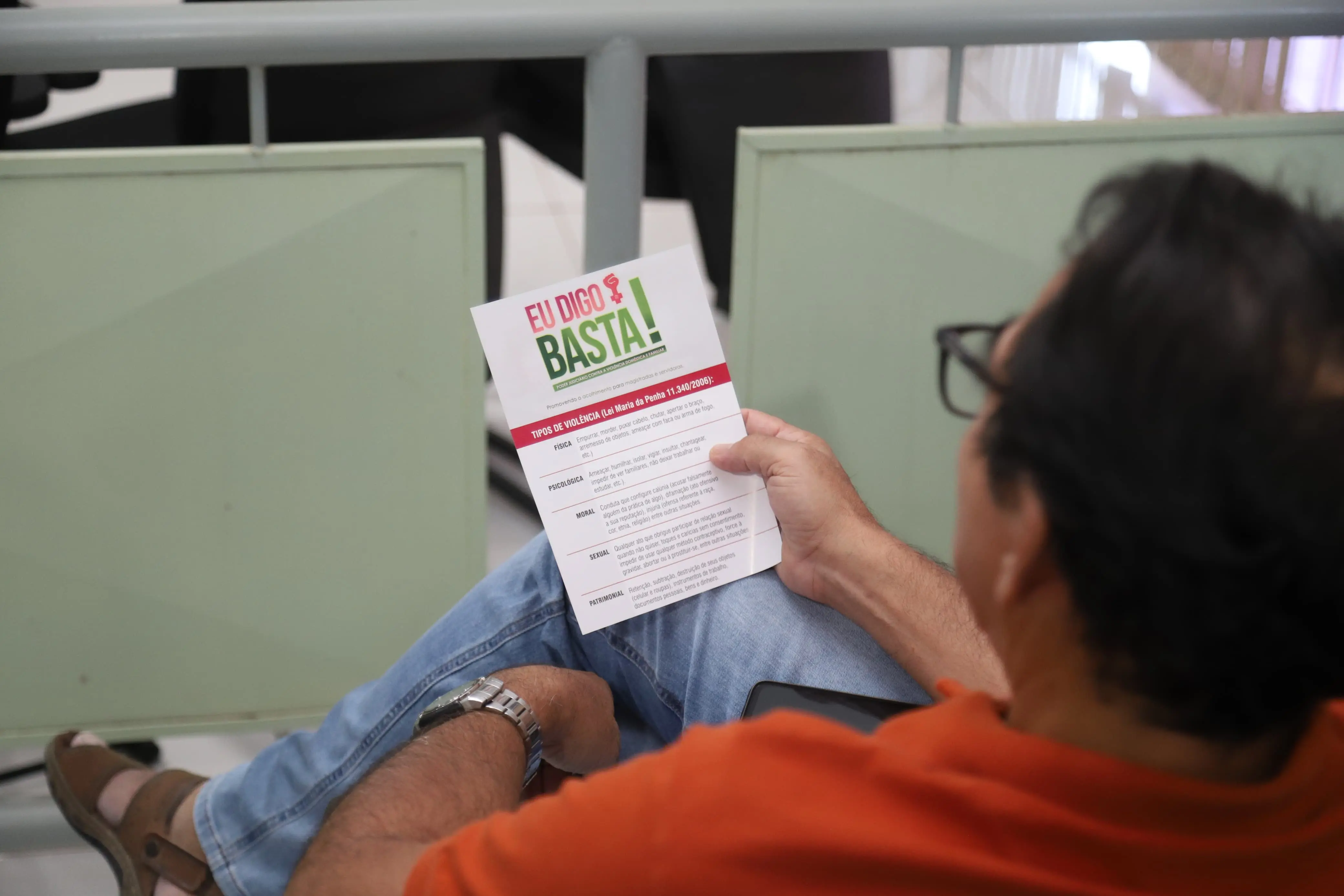 Homem sentado segura um folder da campanha “Eu Digo Basta!”, que lista tipos de violência contra a mulher. Ele veste camisa laranja, calça jeans e sandálias. A imagem destaca o material informativo da Lei Maria da Penha.