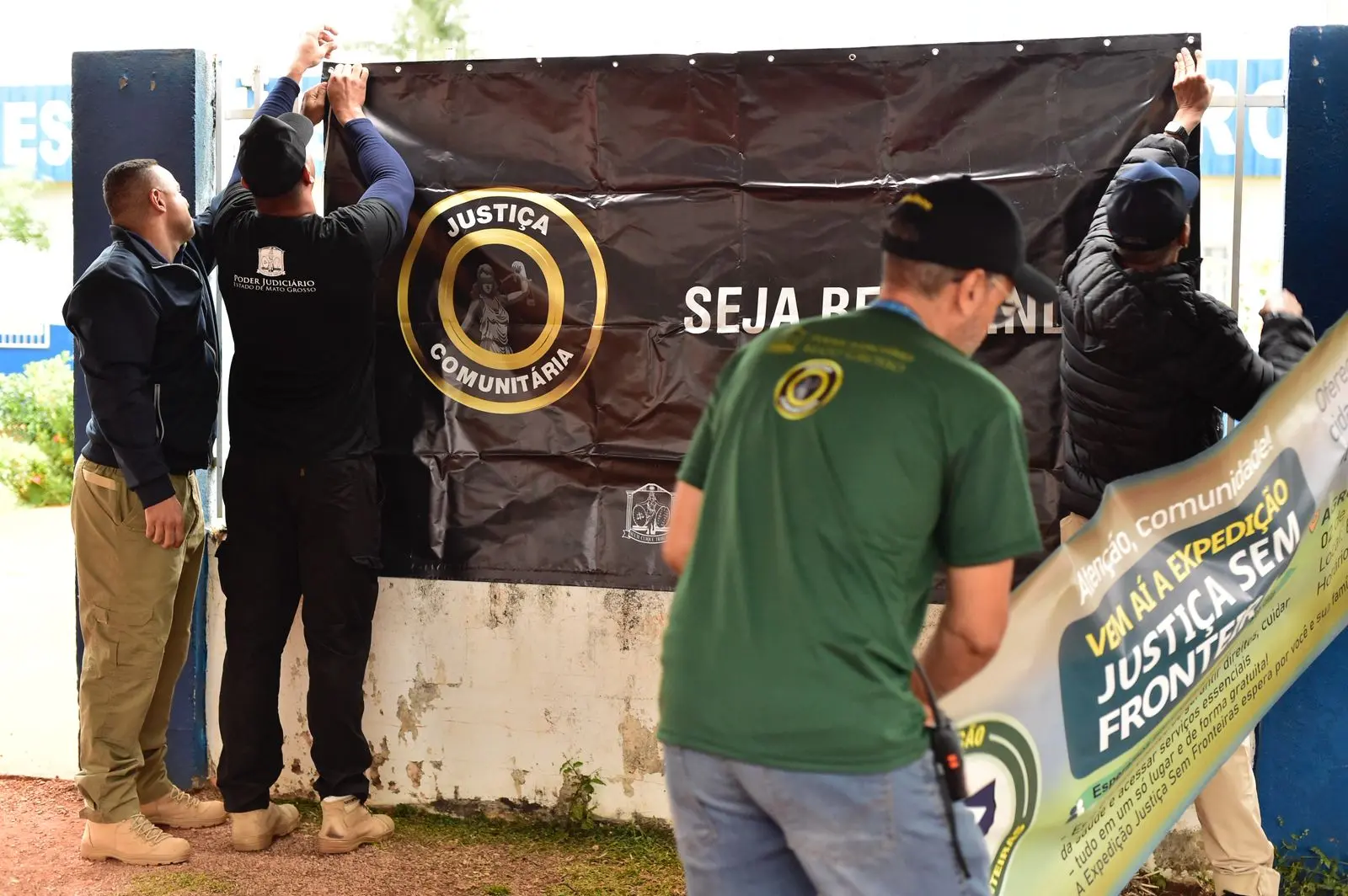 Três homens fixam faixa com os dizeres “Justiça Comunitária – Seja Bem-vindo” na entrada da escola. Um quarto homem segura outro cartaz que anuncia a Expedição Justiça Sem Fronteiras